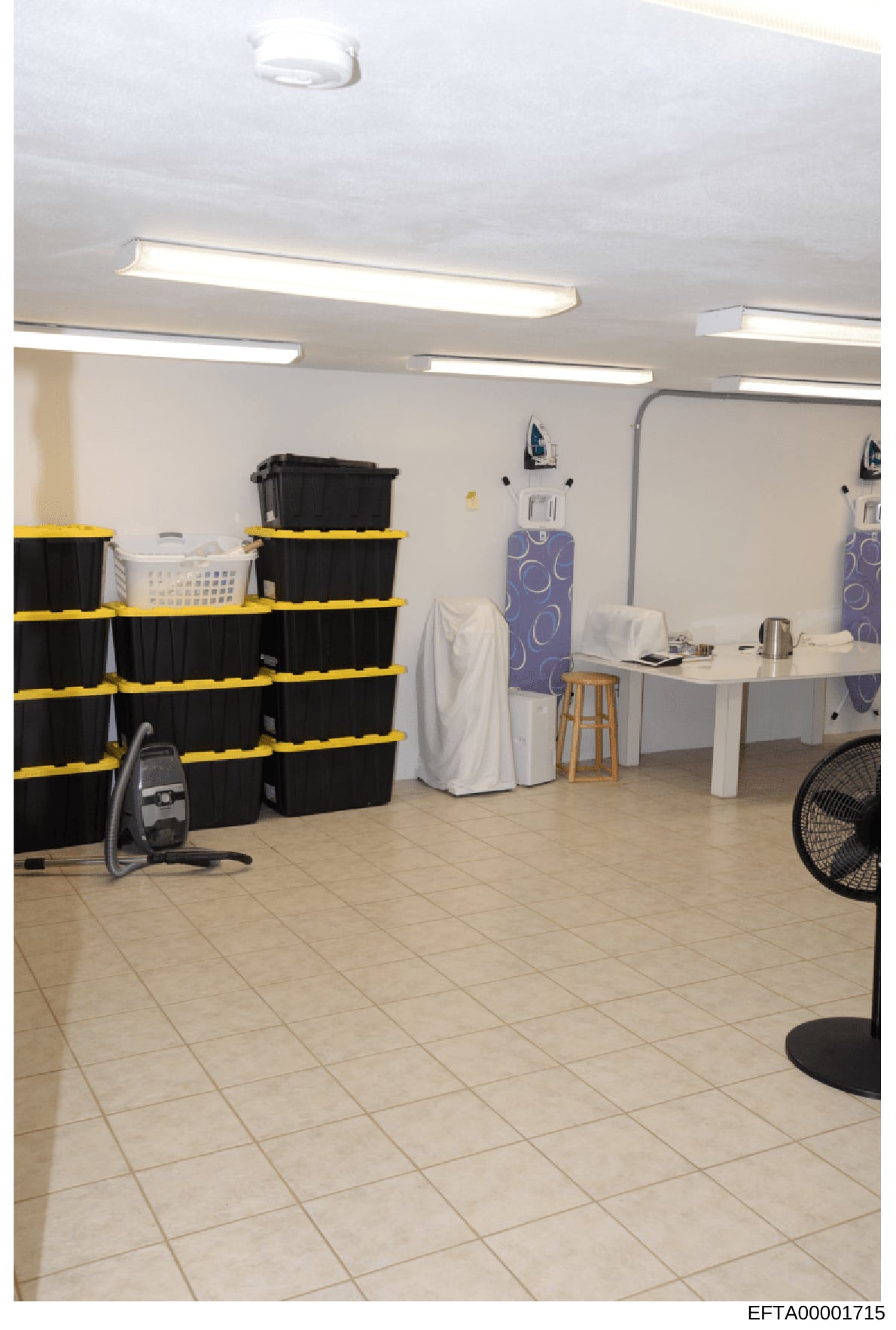 This is an evidence photograph showing the interior of what appears to be a storage, laundry, or utility room. The space contains multiple black plastic storage containers stacked with yellow banding, fluorescent lighting, mirrors, ironing boards, an