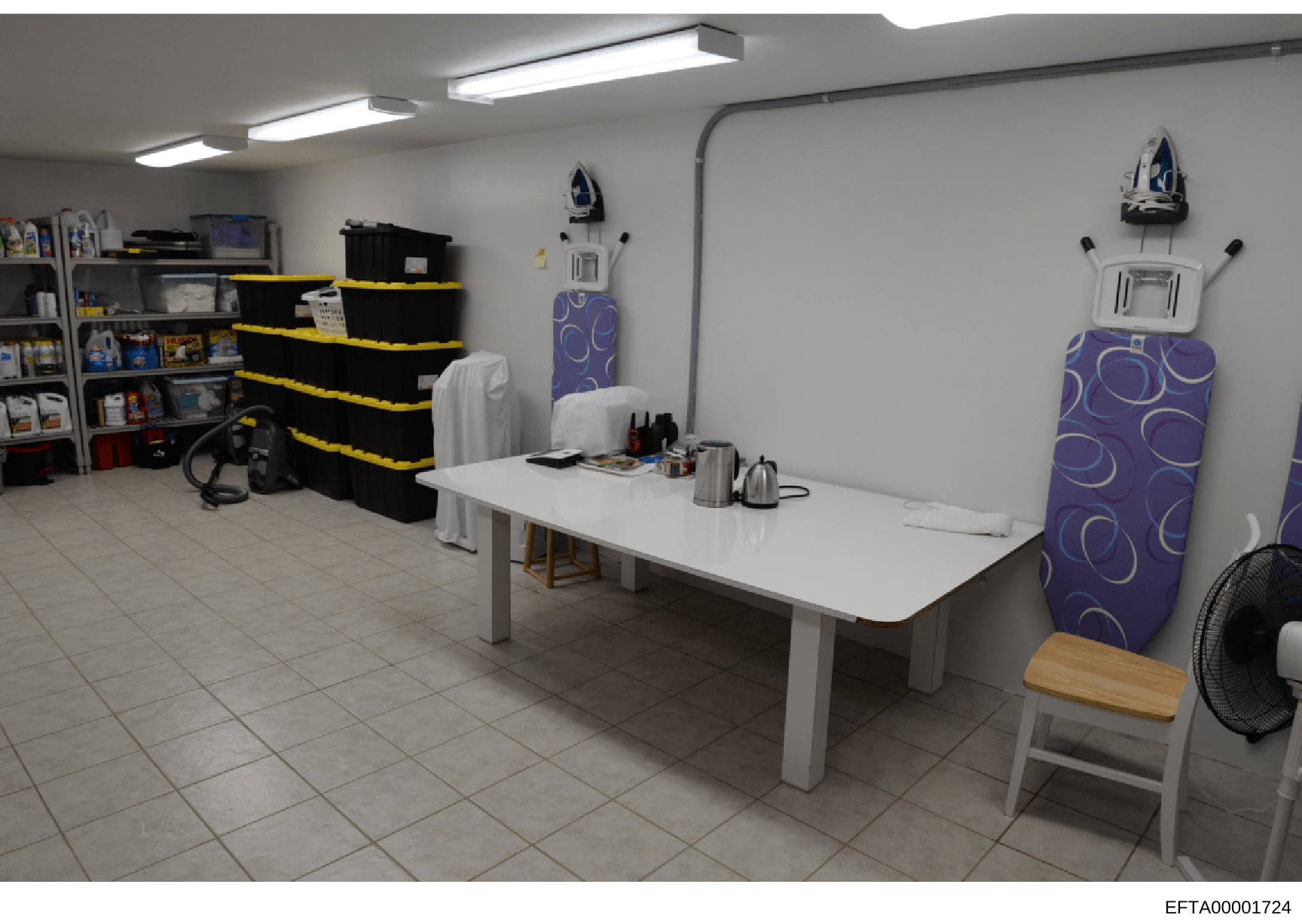 This is a photograph of an interior domestic space showing a utility room or storage area with organized shelving, household items, and appliances. The room appears to be part of a residential property, featuring storage solutions, ironing boards, an