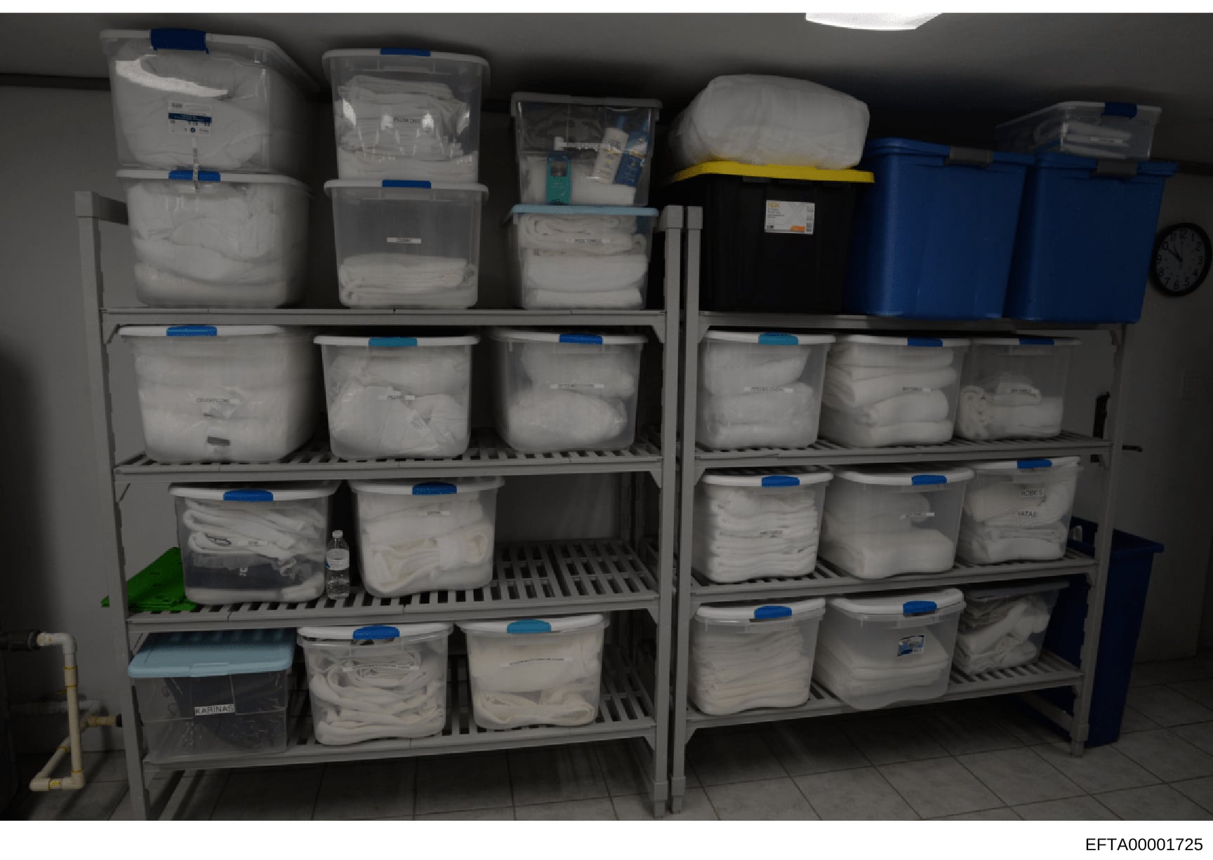 This photograph shows an organized storage facility with multiple shelving units containing numerous plastic storage containers and bins, many labeled with blue tags and labels. The containers appear to be evidence storage or archival storage, consis