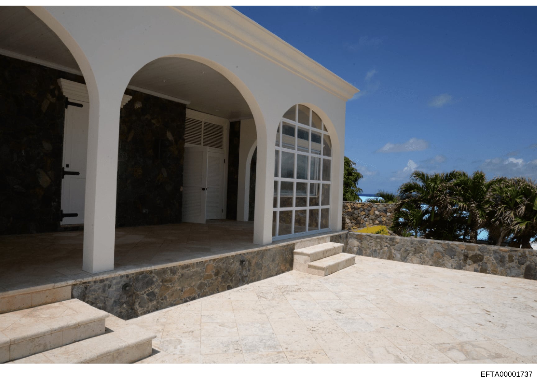 This is a photograph of a contemporary white residential property featuring modern Mediterranean-style architecture with distinctive arched openings. The image shows a covered patio or terrace area with stone accents, coastal landscaping with palm tr