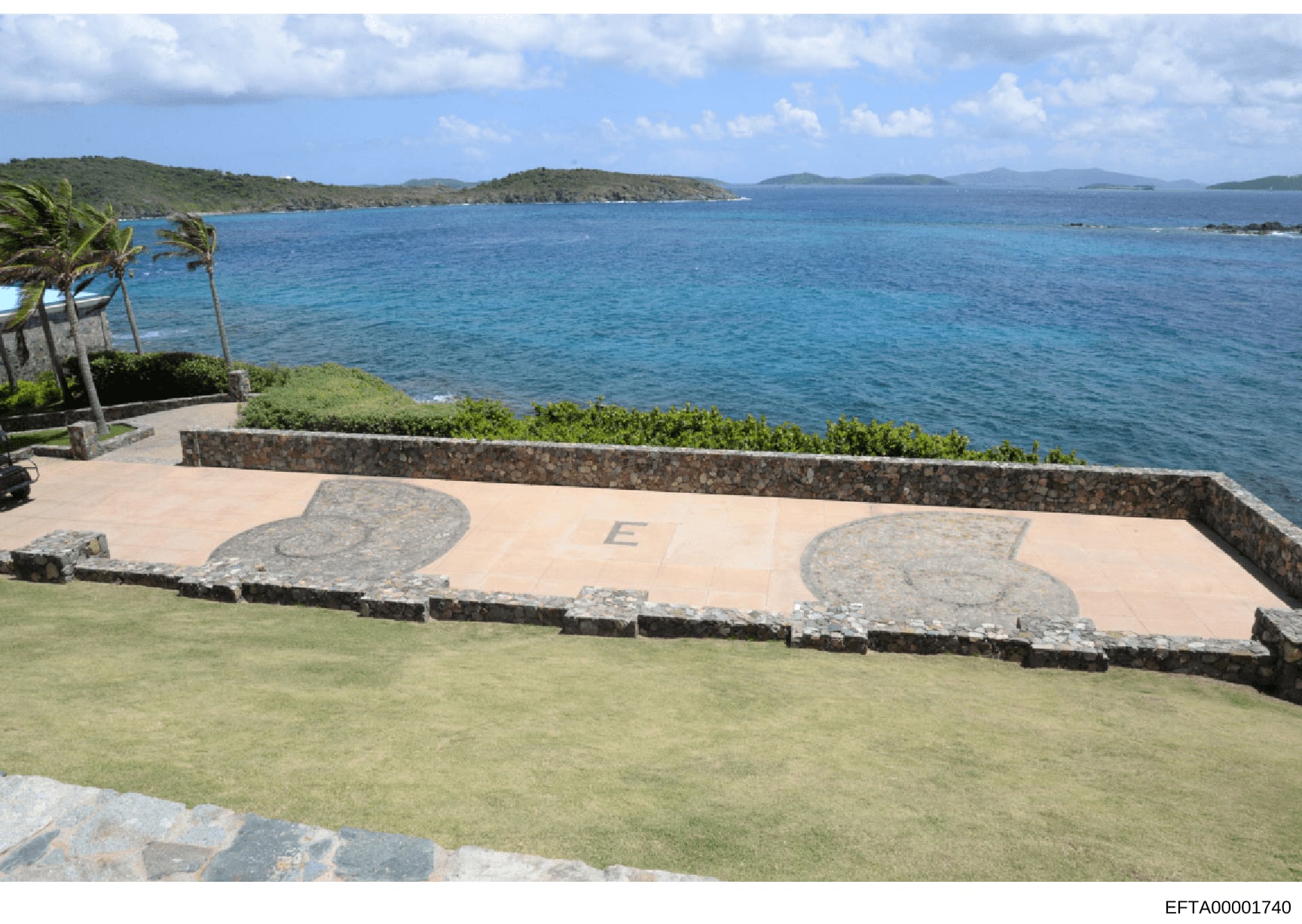 This is a photograph of a scenic oceanfront property featuring a large stone patio with decorative inlaid designs, manicured lawn, and expansive views of turquoise waters and distant islands. The image appears to be documentation of a residential or 