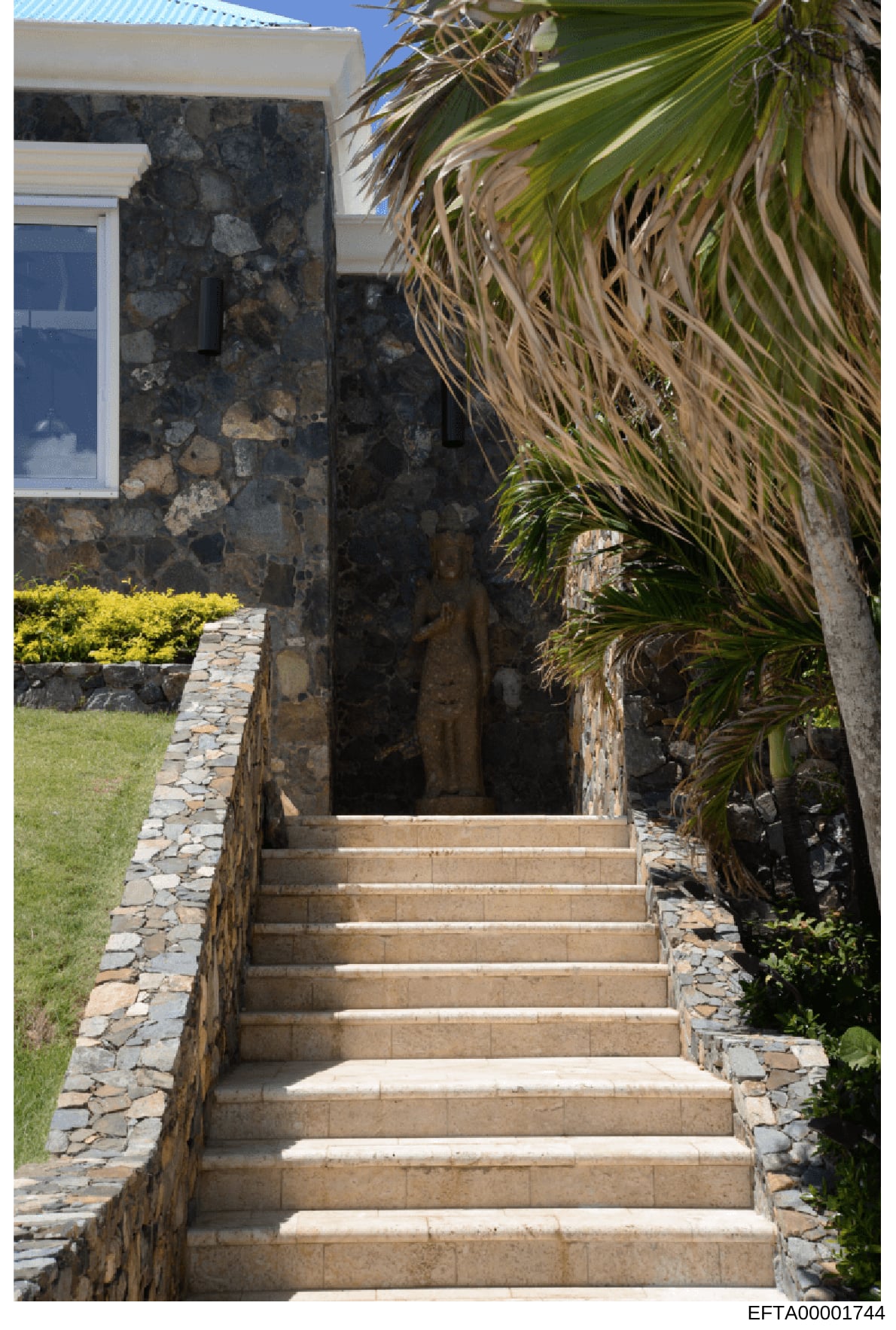 This photograph shows the exterior entrance to a residential property featuring a concrete staircase leading to an elevated entry point. The structure displays stone masonry construction with white trim, tropical vegetation including palm plants, and