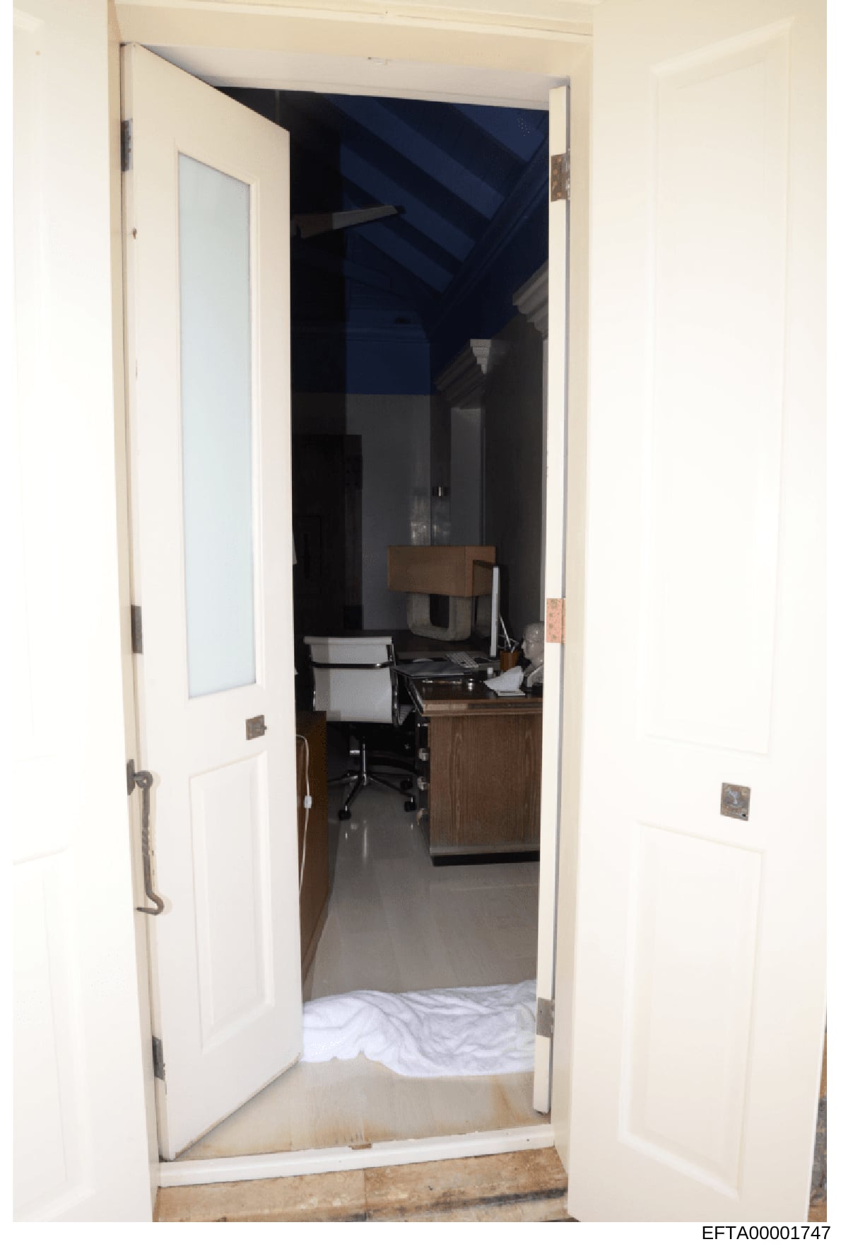 This is a photograph of a room interior taken through an open doorway, showing a workspace with a wooden desk, office chair, and various equipment. The room features cream-colored walls, a dark blue accent wall or structure visible in the background,
