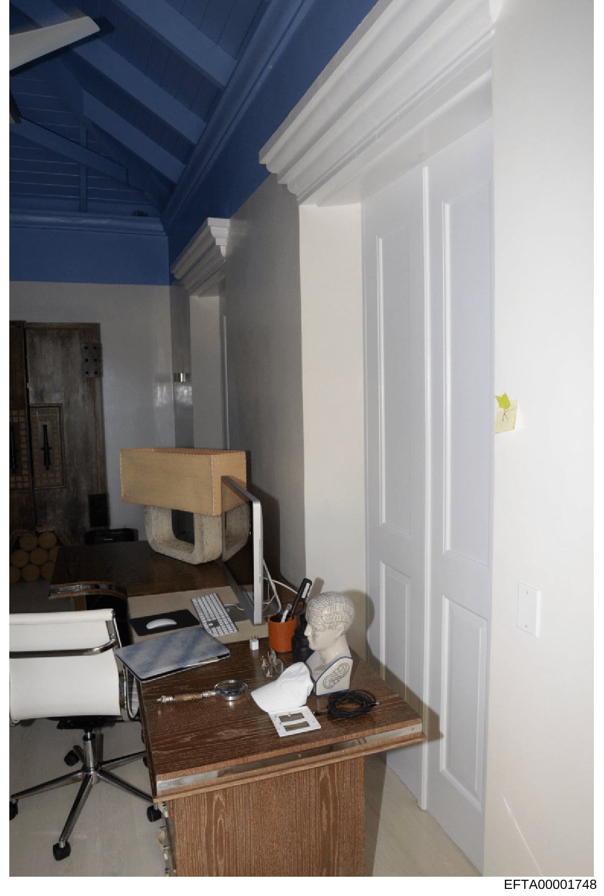 This photograph shows an interior office or study space with a wooden desk, computer setup, office supplies, and decorative elements. The room features white paneled doors, blue ceiling detail, and minimal furnishings. The image appears to be evidenc