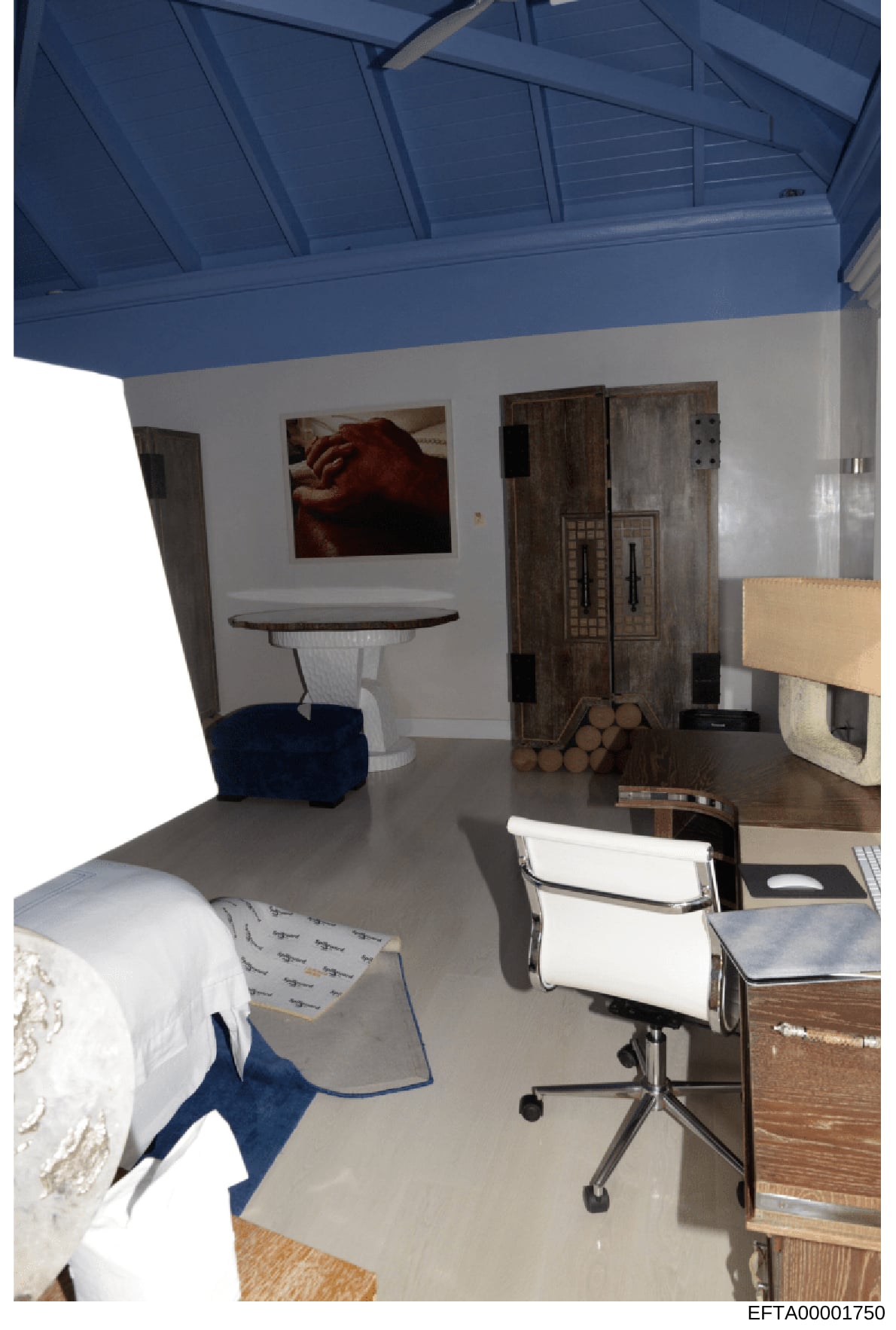 This is a photograph of an interior room, likely from one of Epstein's properties, showing a modern space with a distinctive blue ceiling, white walls, and mixed furnishings including a bed, desk, office chair, round table, and wooden cabinets. The r