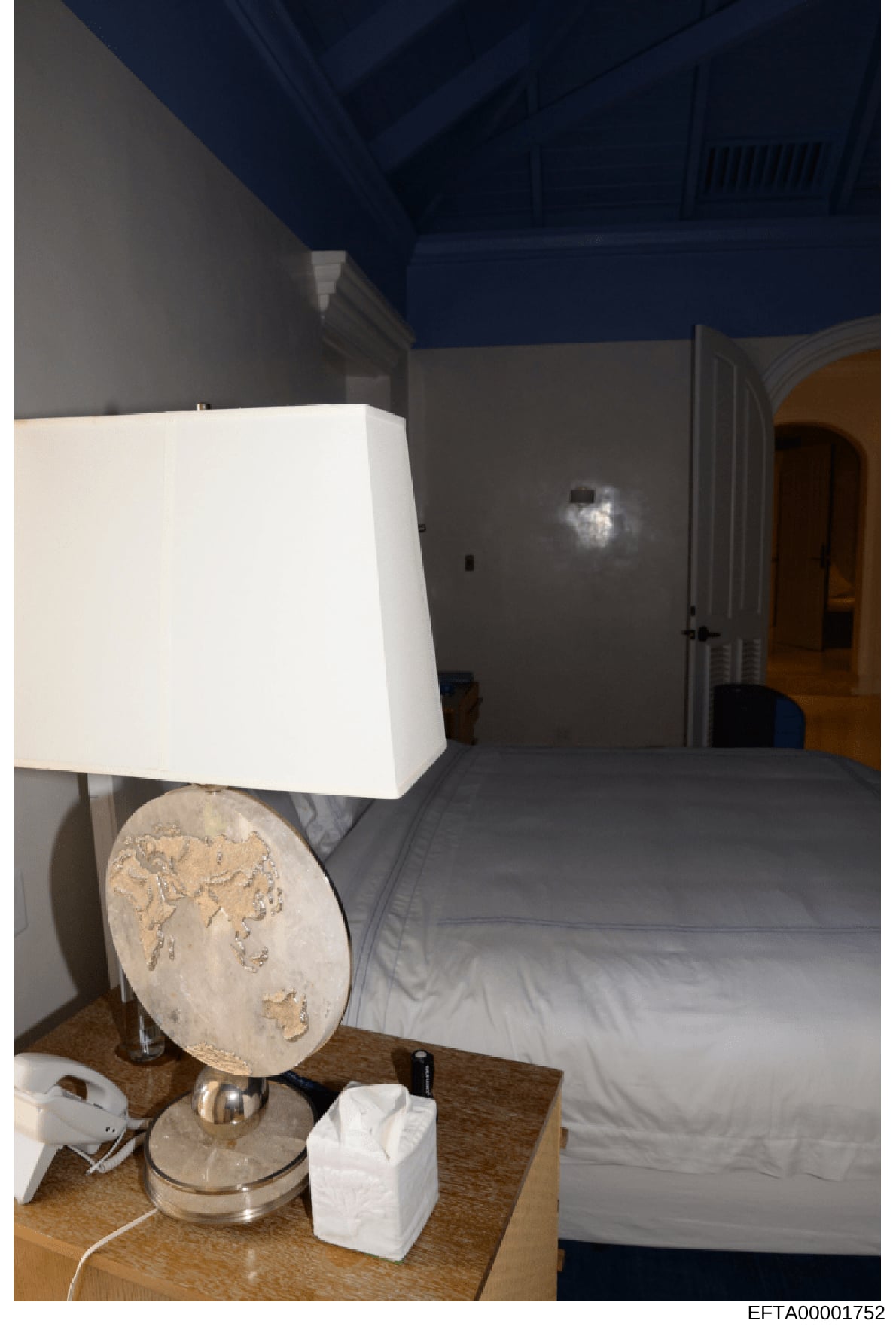Photograph of an interior bedroom space showing a bed with gray bedding, a wooden nightstand with decorative lamp and tissue box, and walls with two-tone paint (gray lower, dark blue upper). The image appears to be documentation of a property's inter