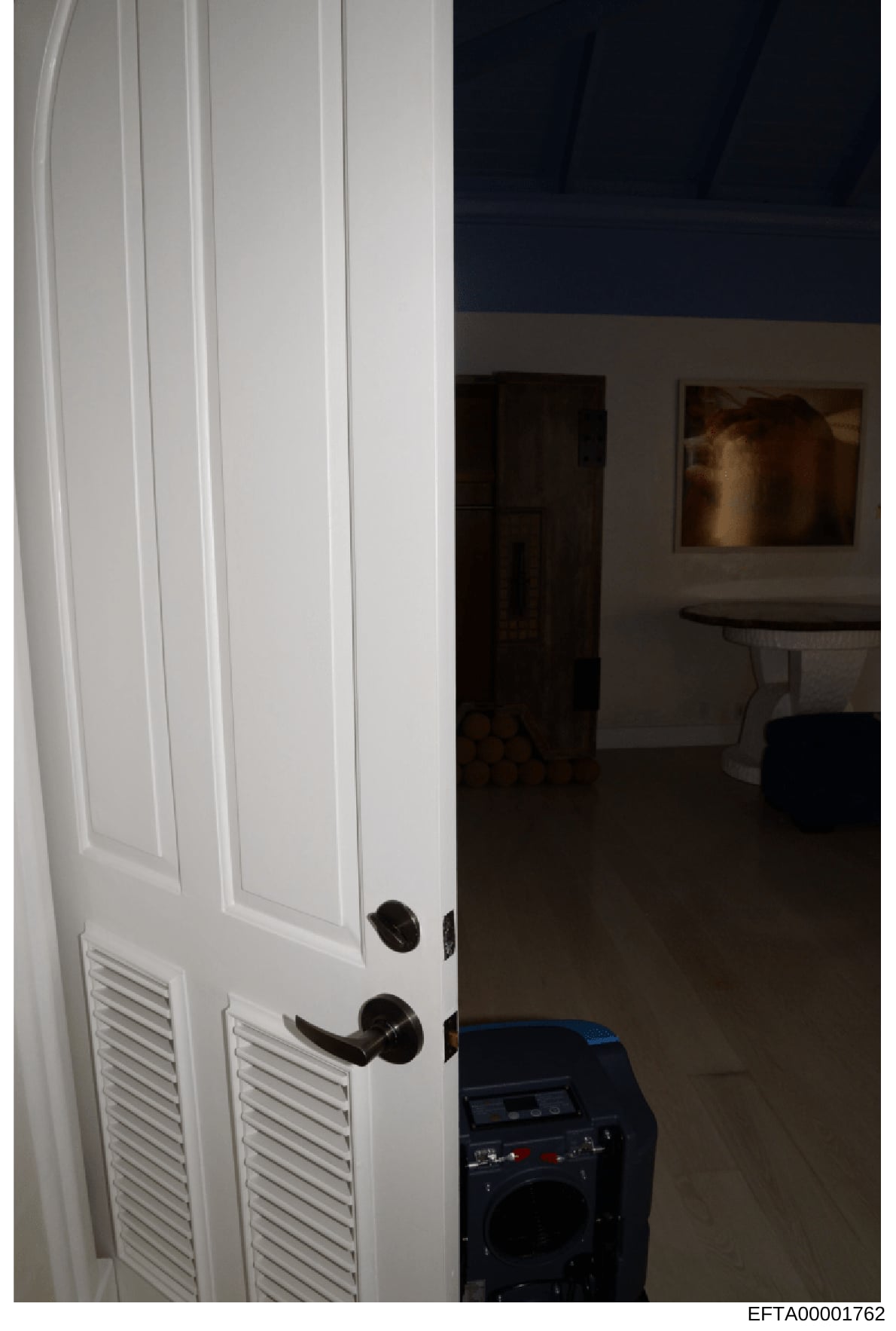 This is an evidence photograph showing an interior doorway with white louvered doors, black hardware, and a view into an adjacent room. The photo captures interior architectural details and furnishings, likely documented as part of property evidence 
