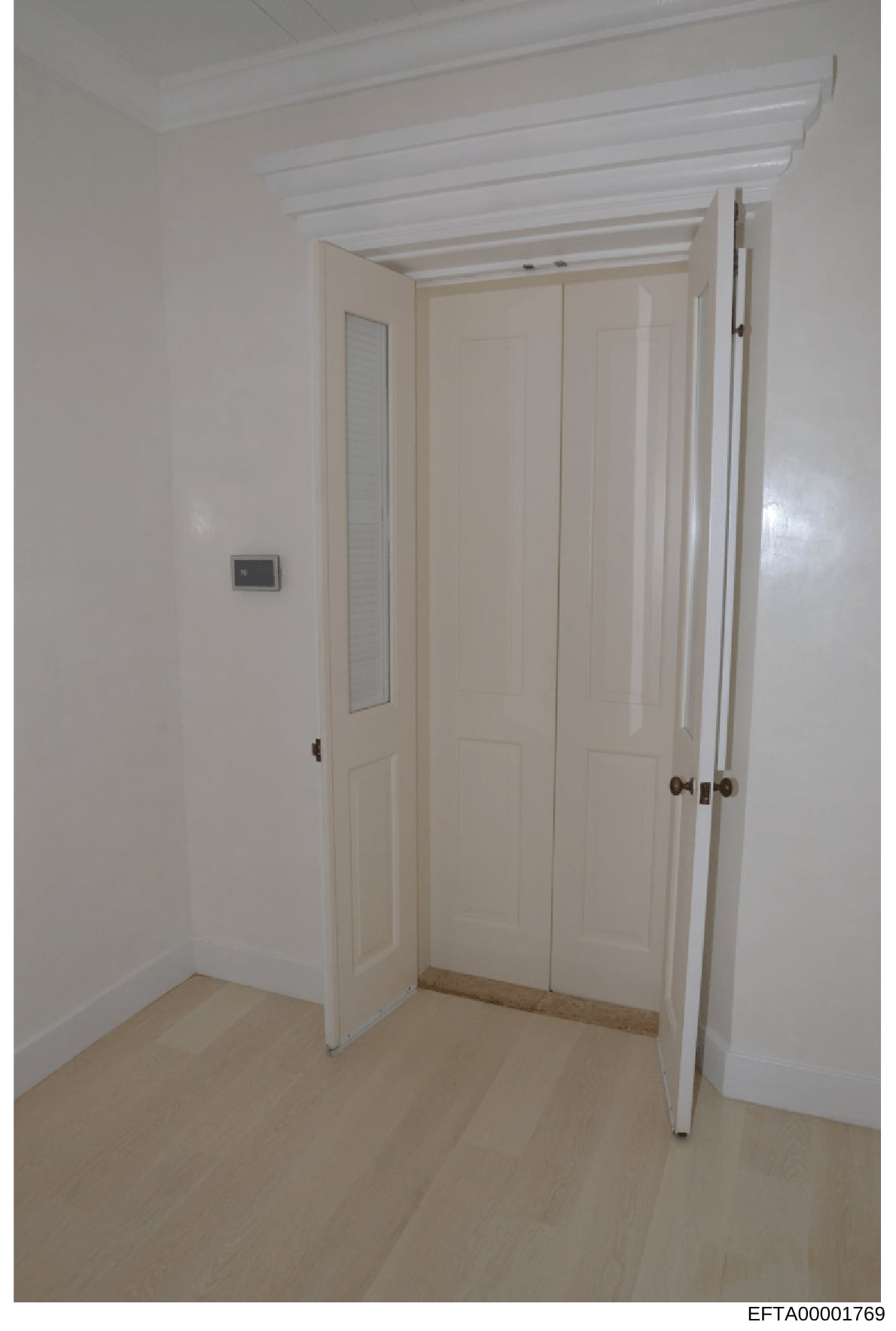 This is a color photograph of an interior hallway or corridor, showing white walls, exposed beam ceiling details, cream-colored closet or wardrobe doors with multiple panels, light wooden flooring, and a small wall-mounted switch or control panel. Th