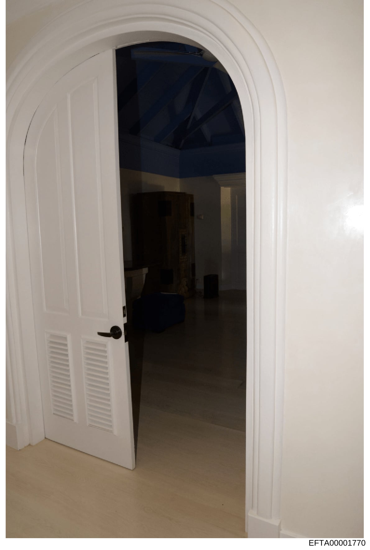 This is a photograph of an interior doorway featuring white trim with an arched molding and a white louvered door. The room beyond the doorway appears to have dark/navy blue walls or ceiling and minimal furnishings. The photograph was taken as part o