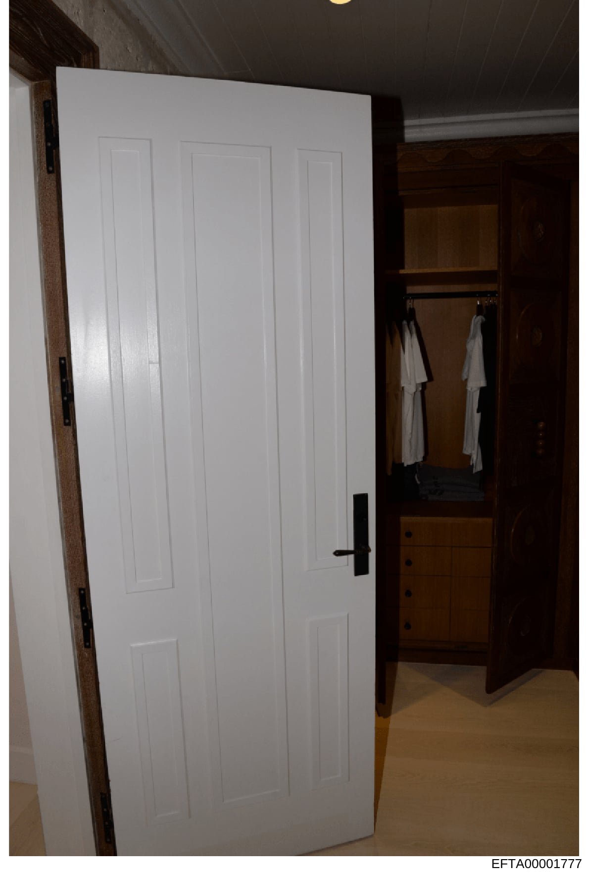 This is an evidence photograph showing the interior of a bedroom closet space with white multi-panel doors, wooden framing, and white garments hanging on a rod inside. The image appears to be part of property documentation from an Epstein investigati