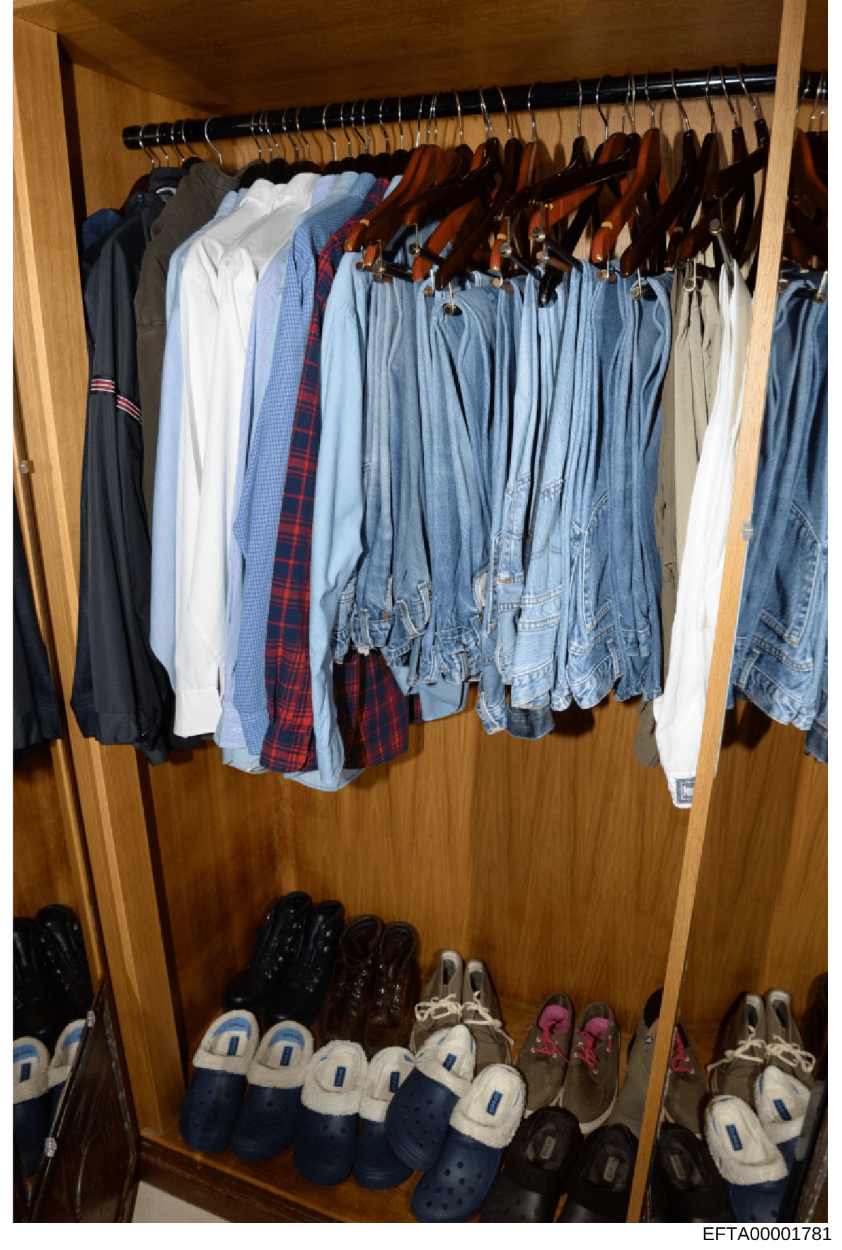 This is a photograph of a wooden closet containing various clothing items on hangers and shoes on the floor. The closet holds a collection of casual clothing including jeans, shirts, sweaters, and jackets in multiple colors, along with casual footwea
