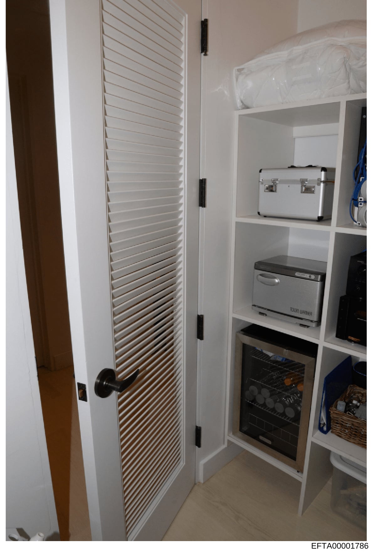 This is a photograph of a utility closet or storage area in one of Epstein's properties, showing built-in white shelving units containing kitchen appliances including a microwave, toaster oven, and other equipment. A louvered door is visible on the l
