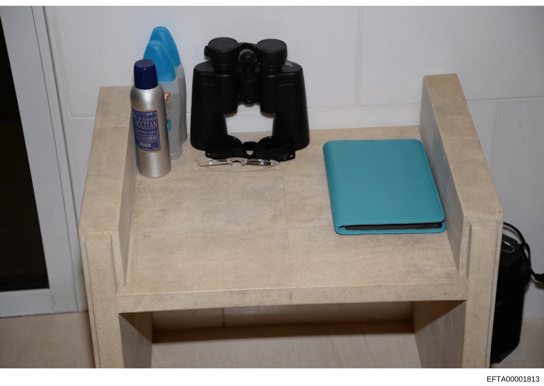 This is an evidence photograph showing a beige/cream-colored nightstand with several personal items arranged on its surface. The items include a metallic spray bottle (appearing to be a hygiene or personal care product), black binoculars, a turquoise