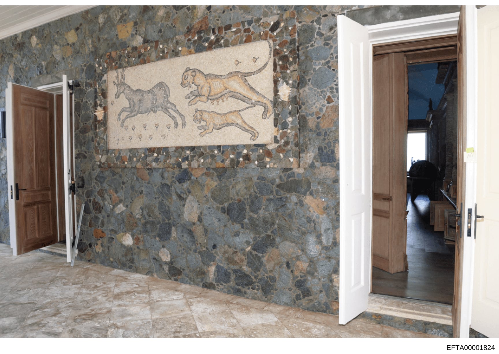 This photograph documents an interior hallway or corridor featuring a decorative mosaic tile wall and classical artwork. The space displays a sophisticated multi-colored stone or terrazzo wall with embedded tiles in earth tones, along with a framed m