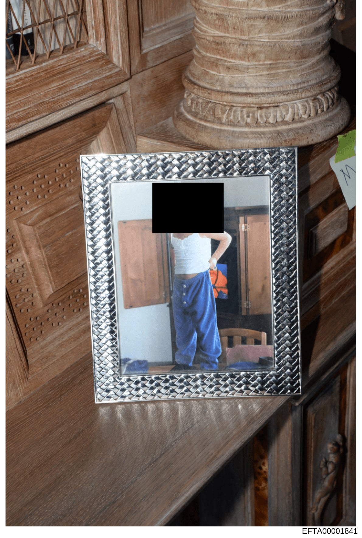 This is an evidence photograph showing a framed picture discovered during what appears to be an interior search of a property. The photograph itself is partially redacted (face obscured with black redaction bar). The frame is ornate with a metallic b