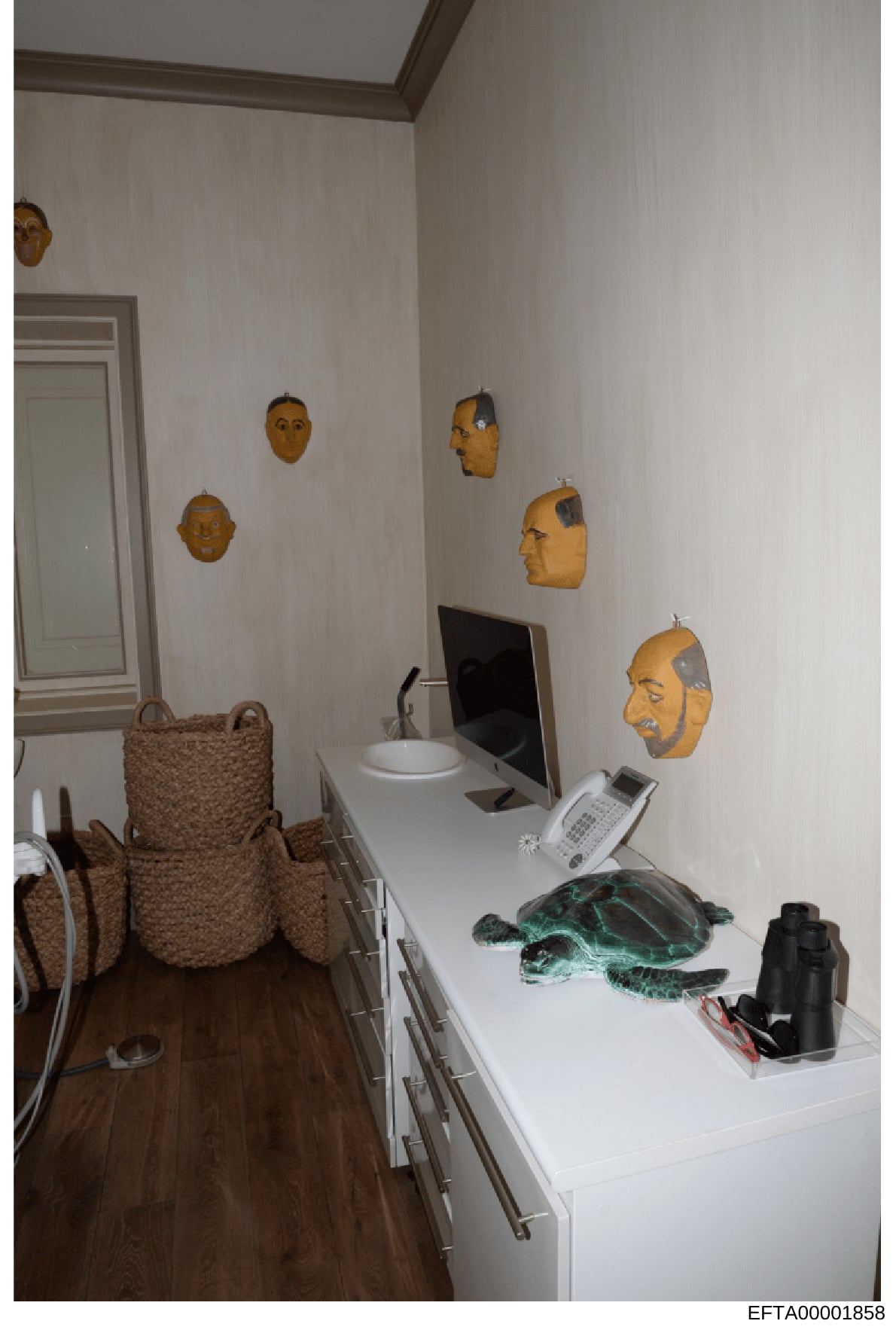 This photograph depicts an interior room, likely an office or study, displaying decorative wooden or bronze masks on the walls and various objects on a white desk. The image appears to be evidence documentation from the Epstein investigation, showing