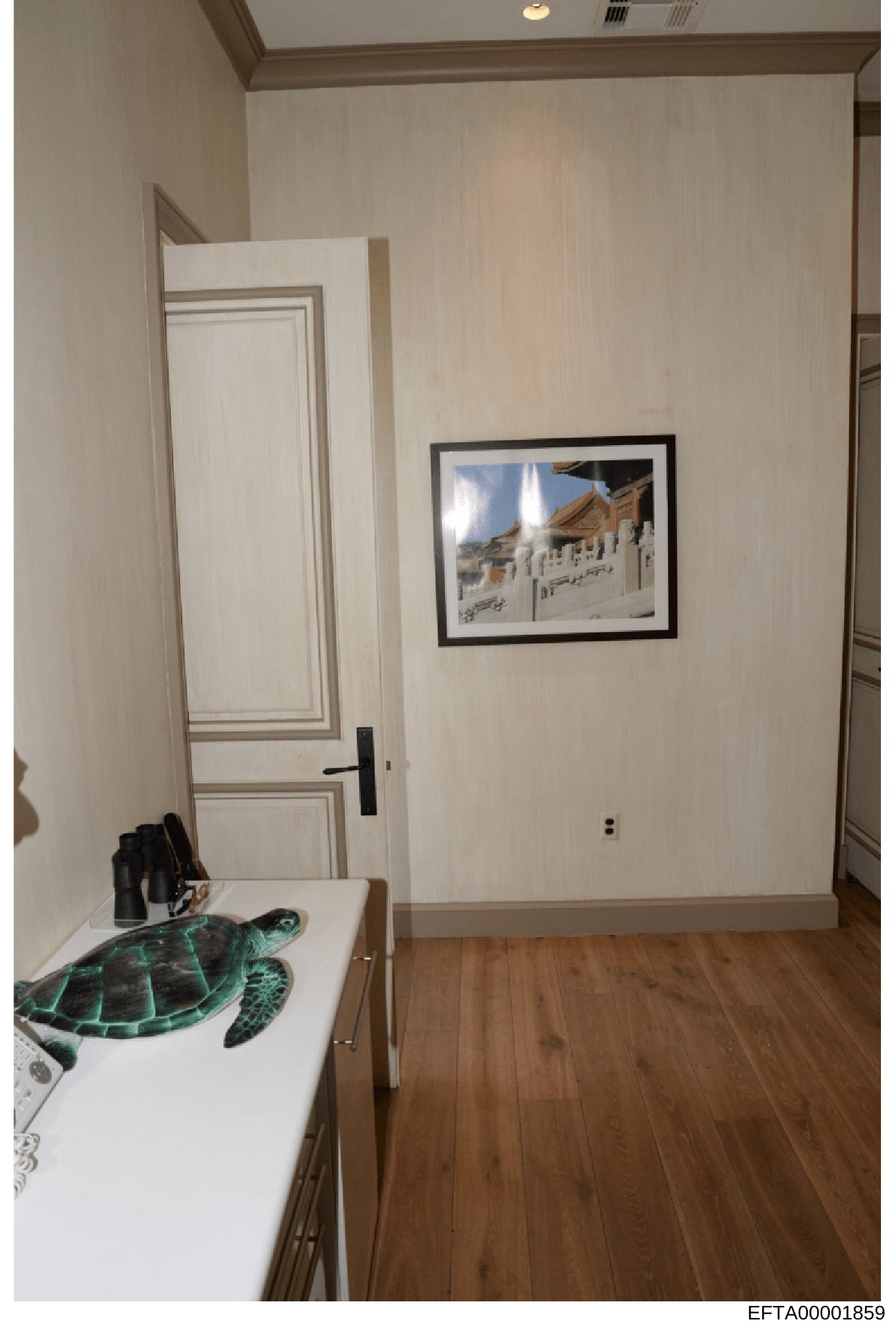 This is a photograph of an interior bedroom space, likely from one of Epstein's properties. The room features cream-colored walls, hardwood flooring, a white dresser with a sea turtle decorative item, and a framed photograph of an Asian architectural