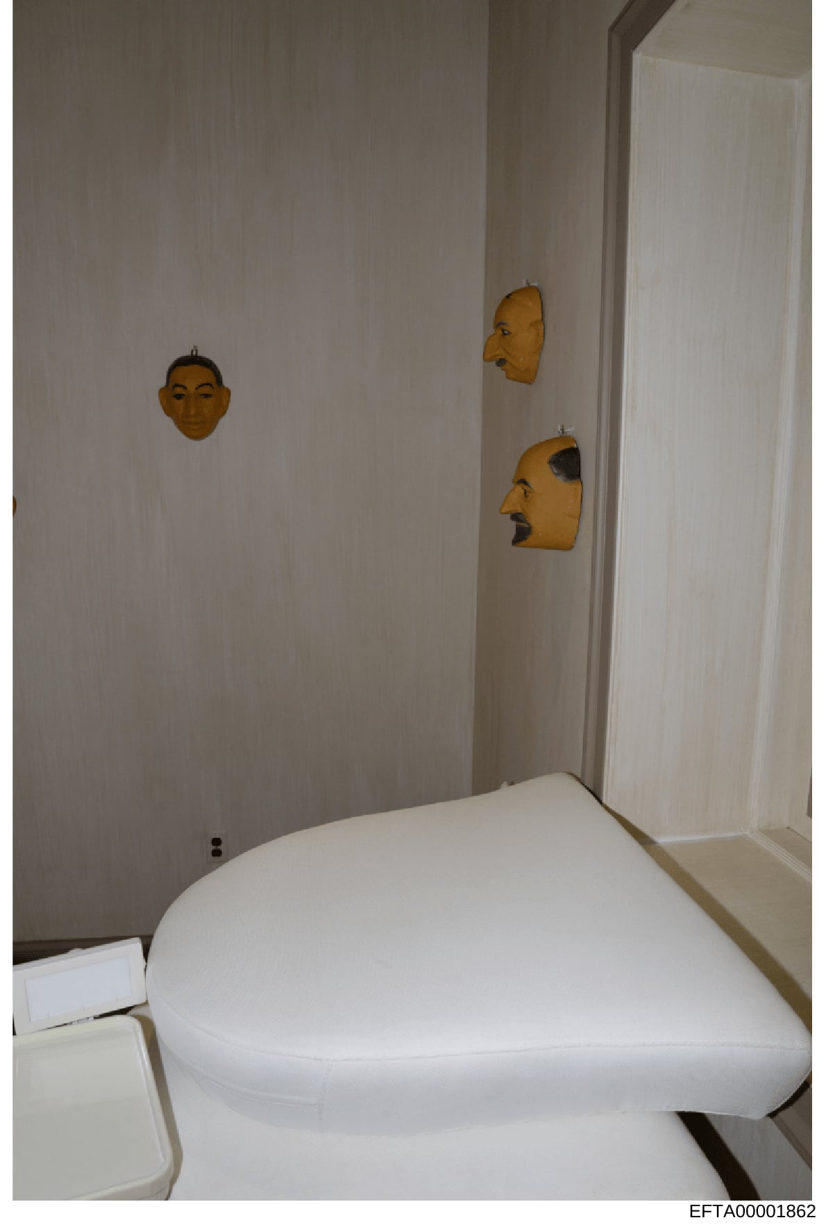 This is an evidence photograph from an Epstein property showing a bathroom interior. The room features three decorative wooden masks hanging on light-colored walls above a white toilet. The masks appear to be carved wooden art pieces in varying style