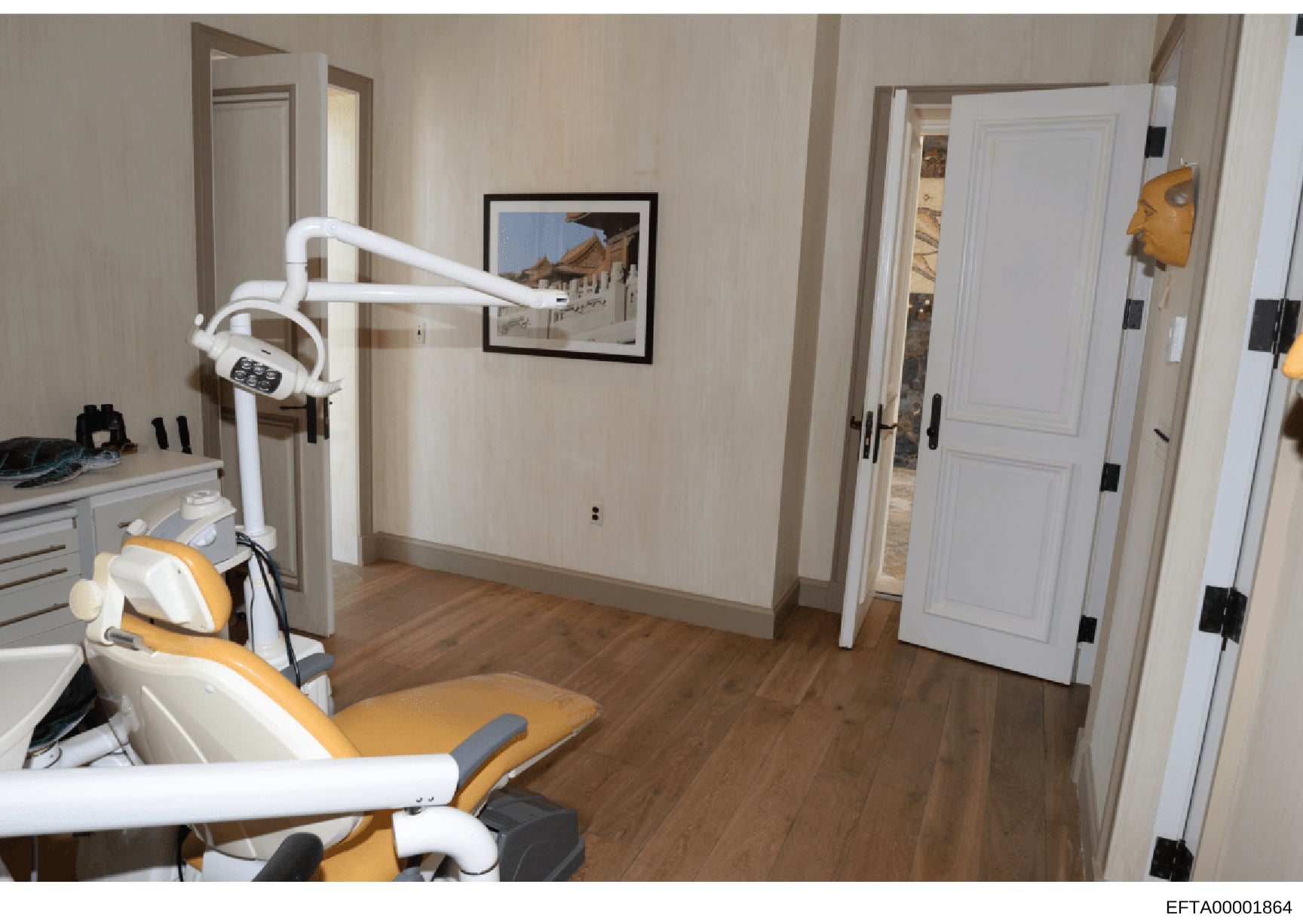 This photograph documents a dental treatment room featuring modern clinical equipment, including a dental chair with yellow upholstery, overhead surgical lighting, and cabinetry. The room has cream-colored walls, white trim, wooden flooring, and fram