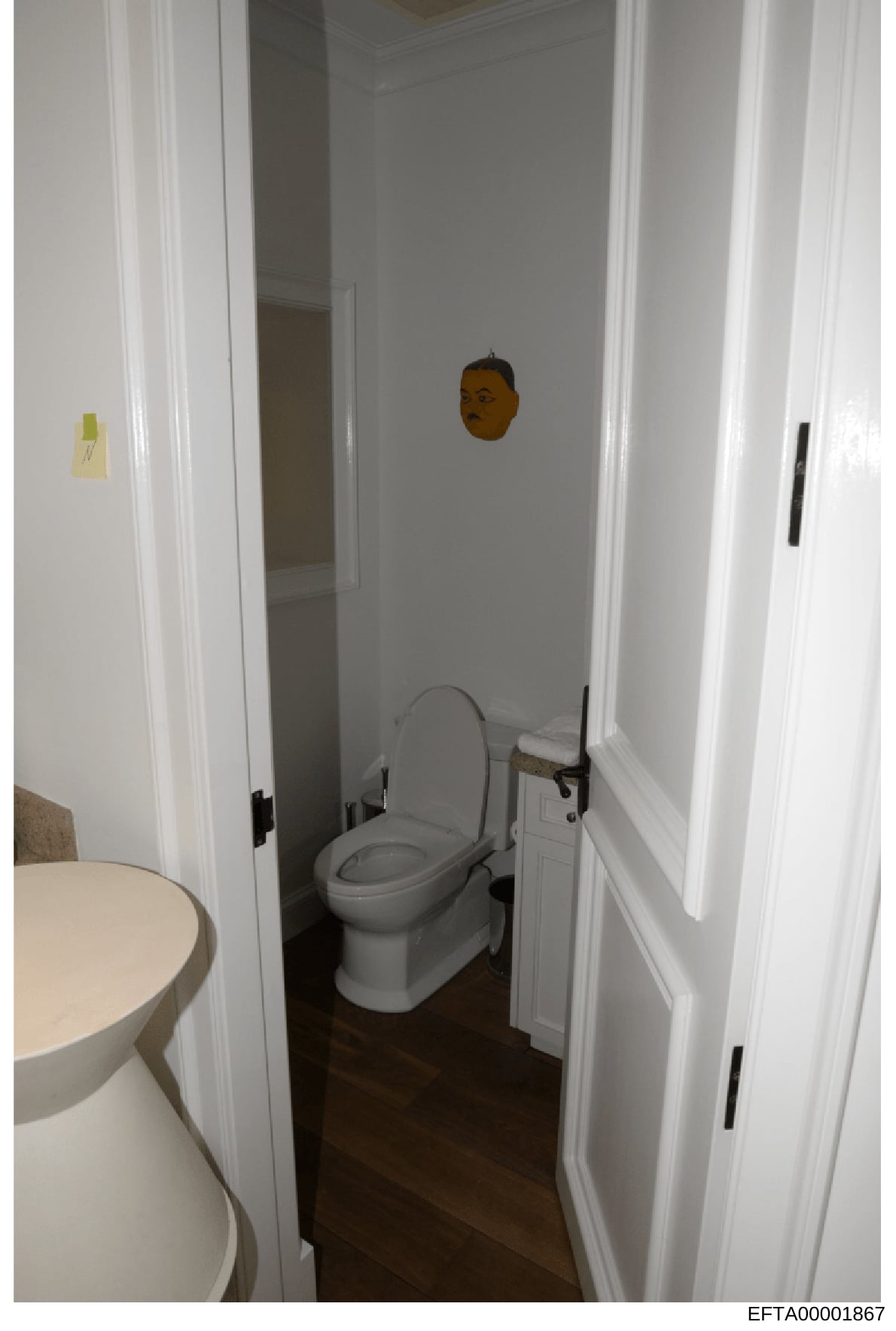 This is a photograph of a bathroom interior showing a toilet, sink, and white-painted walls with dark flooring. The image appears to be evidence documentation from a property inspection or seizure, with evidence markers visible on the walls. The Bate