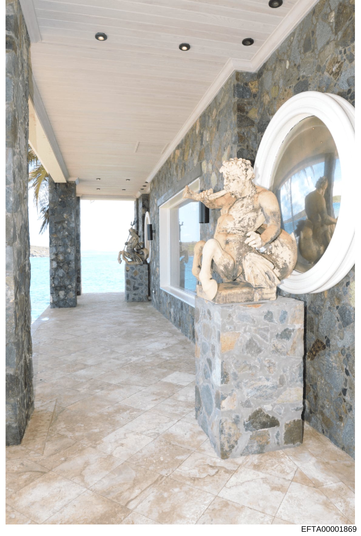 This is an interior photograph of a luxurious coastal property showing a long hallway with stone walls, high white ceiling with recessed lighting, and decorative classical sculptures. The hallway opens to ocean views, featuring ornamental urns and cl