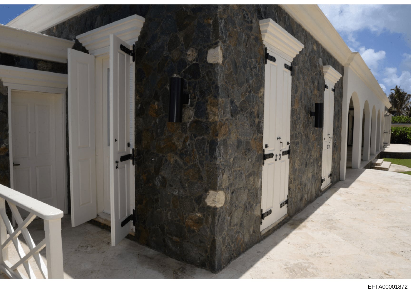 This is a photograph of an outdoor structure featuring dark stone walls with white-trimmed doors and hardware. The building appears to be a poolhouse, cabana, or similar outbuilding at a residential property. The image shows the architectural details