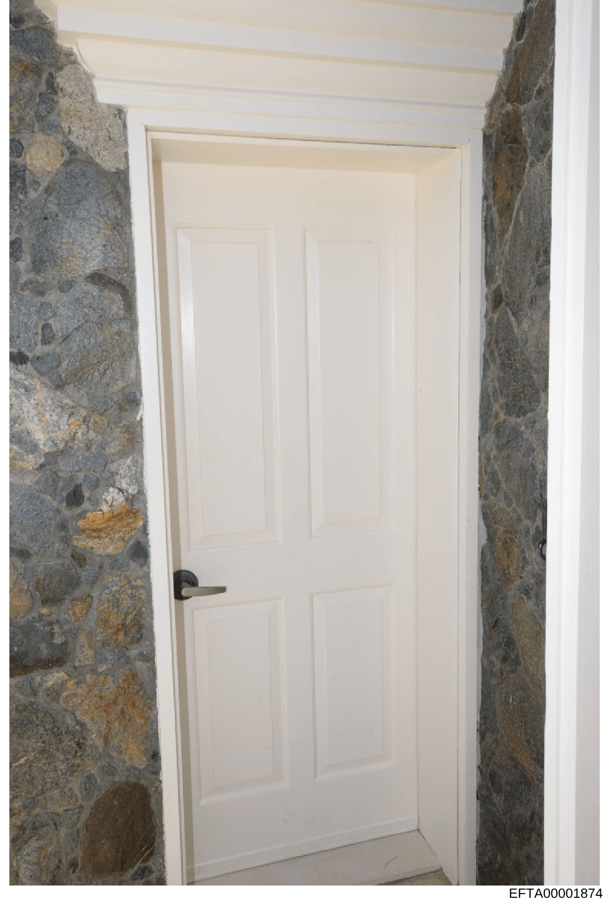 This photograph shows an interior passageway or corridor featuring rustic stone walls in gray and tan tones with a white six-panel door at the far end. The image appears to be documentation of interior architectural features or property conditions, l