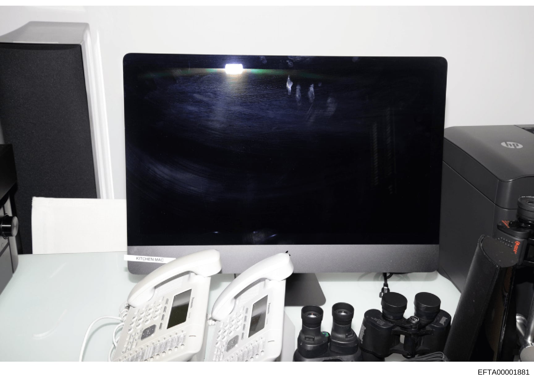 This photograph shows a desktop computer setup labeled 'KITCHEN MAC' with what appears to be surveillance or monitoring equipment, including two white IP phones/devices and multiple camera lenses. The image documents the physical equipment configurat