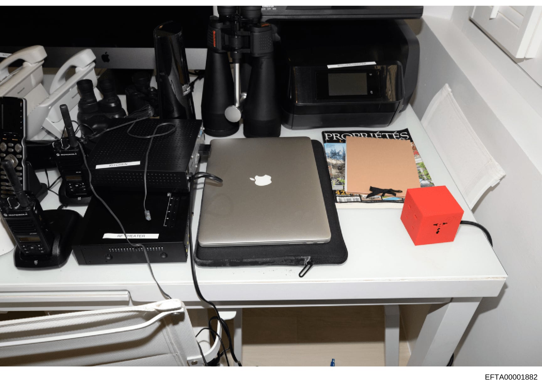 This is a photograph of a white desk containing various electronic equipment, surveillance devices, a laptop computer, and printed materials including what appears to be a magazine or publication labeled 'PROPHETES'. The image documents the physical 
