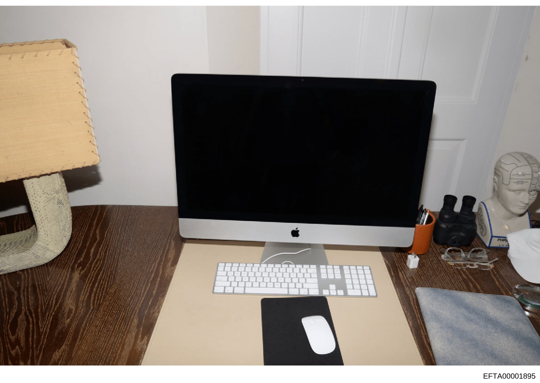 This is an evidence photograph showing a residential or office desk setup featuring an Apple iMac computer with peripherals. The image documents the physical environment and equipment, likely seized or photographed during the investigation. The desk 