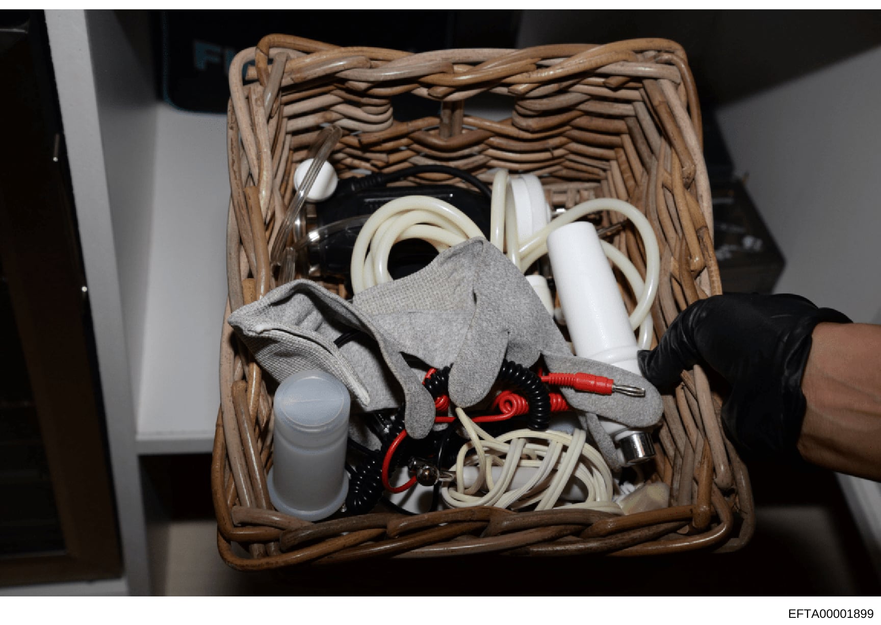 This is an evidence photograph showing the contents of a wicker basket seized during the investigation. The basket contains various household items including cleaning supplies, cords, cables, gloves, and other miscellaneous objects. The photograph wa
