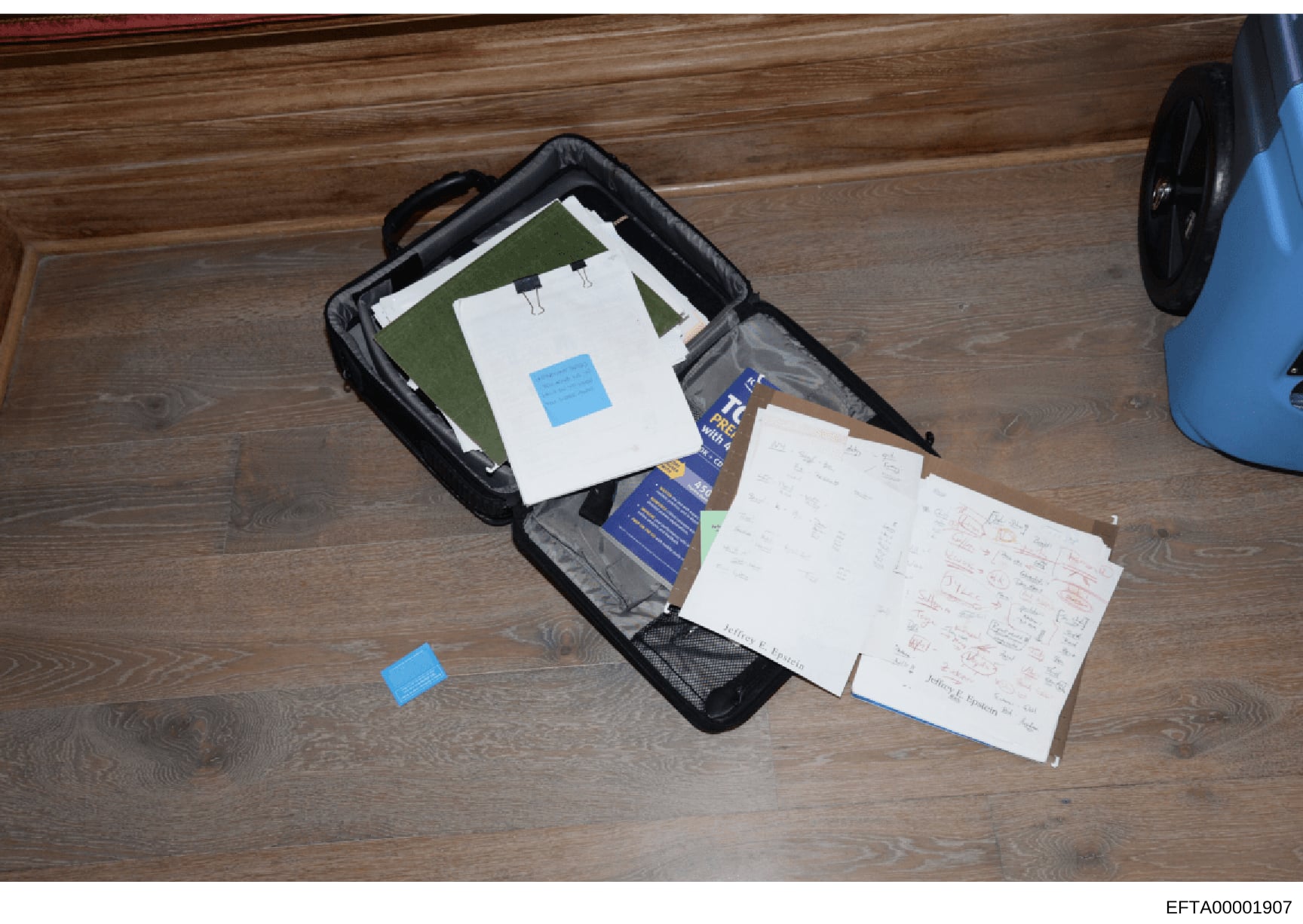 This is an evidence photograph showing a black travel bag or briefcase opened on a wooden floor, containing various documents, papers with handwritten notes, a blue instructional booklet, and what appears to be architectural or planning diagrams. The