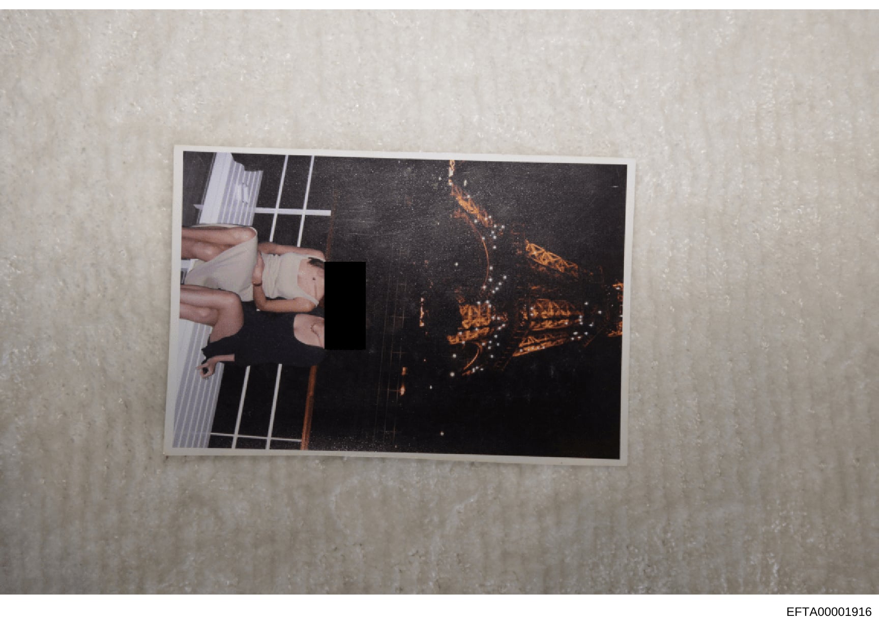 This is an evidence photograph containing a printed image placed on a light-colored surface. The photograph shows a partially visible scene with significant redaction (black bar) obscuring the central content. The left side shows what appears to be h