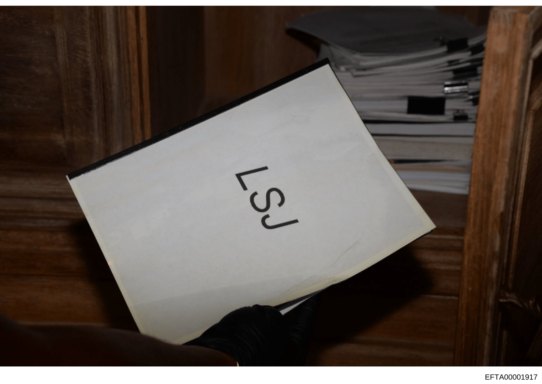This is a photograph of a document box or evidence container labeled 'LSJ' positioned on a wooden surface, with stacked files or documents visible in the background. The image appears to be taken during evidence collection or storage documentation, w