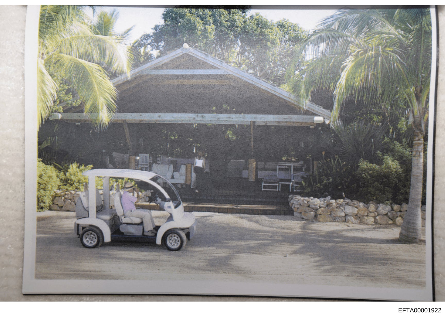 This is a photograph of a residential property featuring a large covered structure with a peaked roof, surrounded by tropical vegetation including palm trees. A white golf cart is parked in the foreground on a paved driveway. The image appears to be 