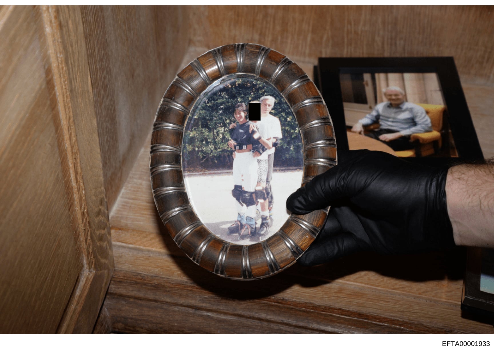 This is an evidence photograph showing a framed oval portrait being held by an investigator wearing black gloves. The framed photo depicts two young people outdoors with foliage in the background, and one face is redacted with a black block. In the b