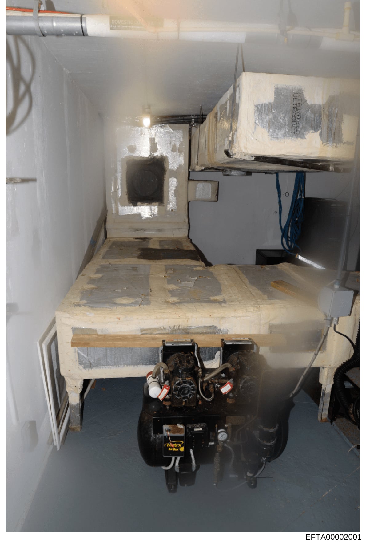 This is a photograph of a confined underground space containing HVAC/mechanical equipment, electrical systems, and storage. The room appears to show a basement or sub-level utility area with exposed wiring, ductwork, and what appears to be a furnace