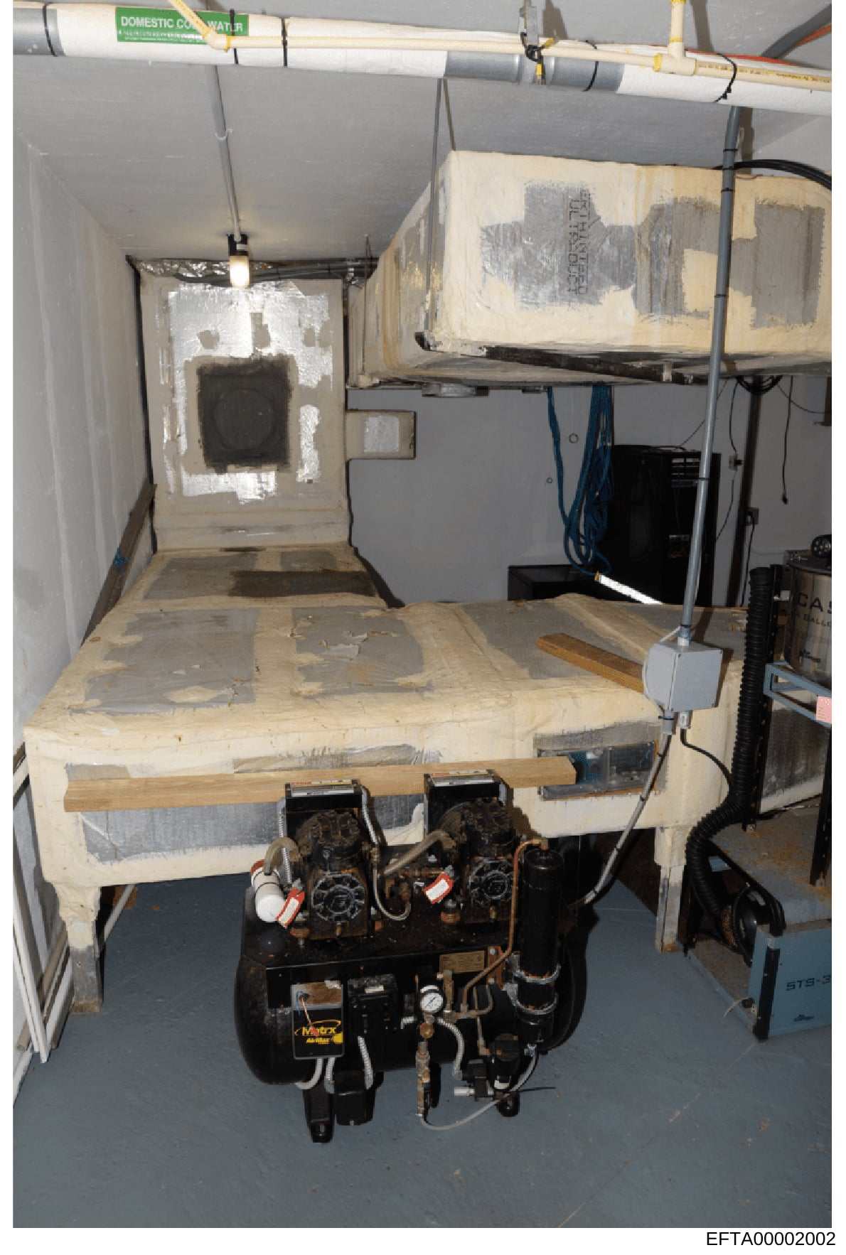 This photograph documents the interior of a basement or mechanical room, showing HVAC equipment, ductwork, and utility installations. The space appears to be under renovation or investigation, with plastic sheeting covering surfaces and walls. The im