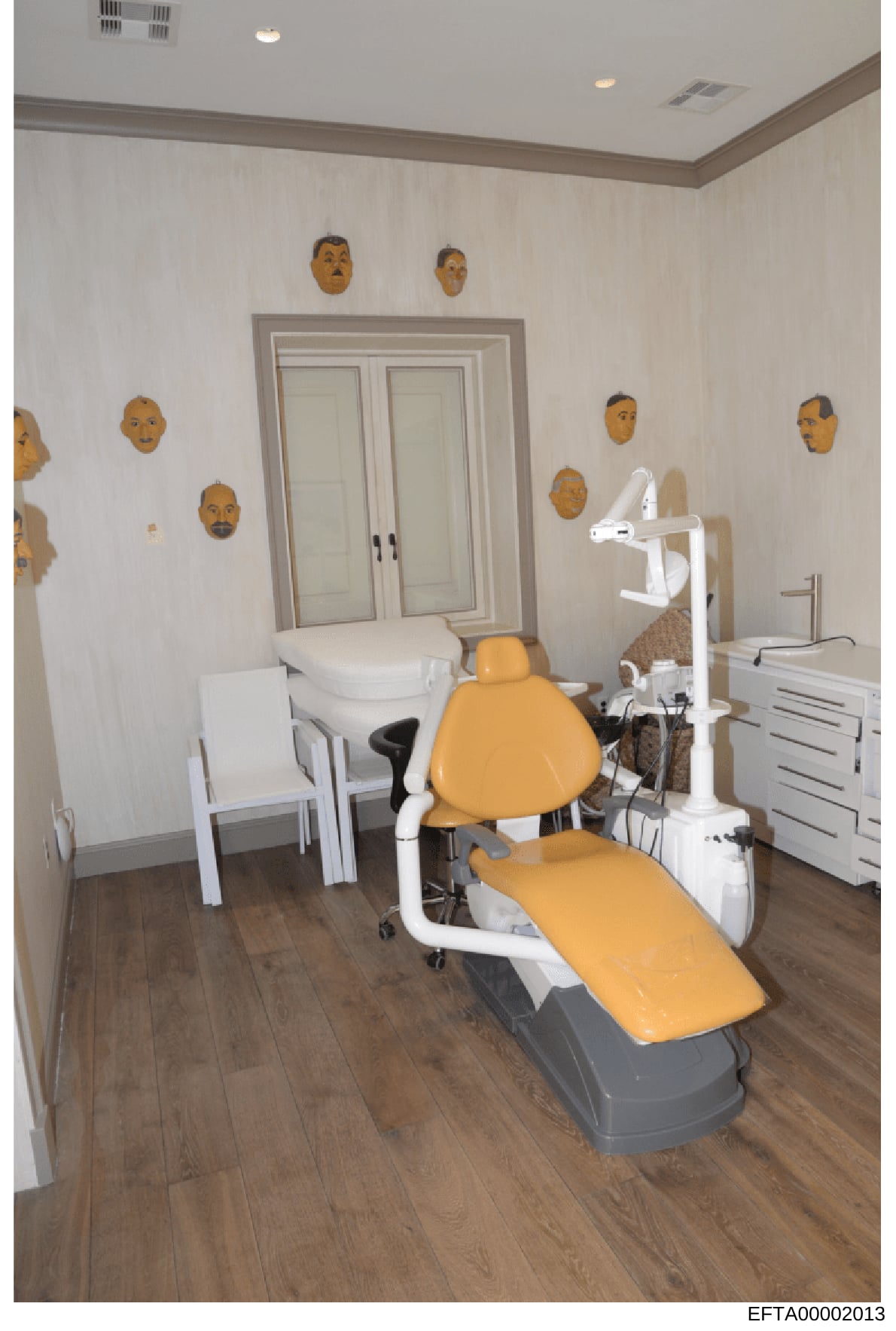 This is a photograph of a modern dental or medical treatment room featuring a yellow dental chair, overhead lighting equipment, and white cabinetry. The walls are decorated with approximately 10-12 decorative wooden face masks or sculptures in golden