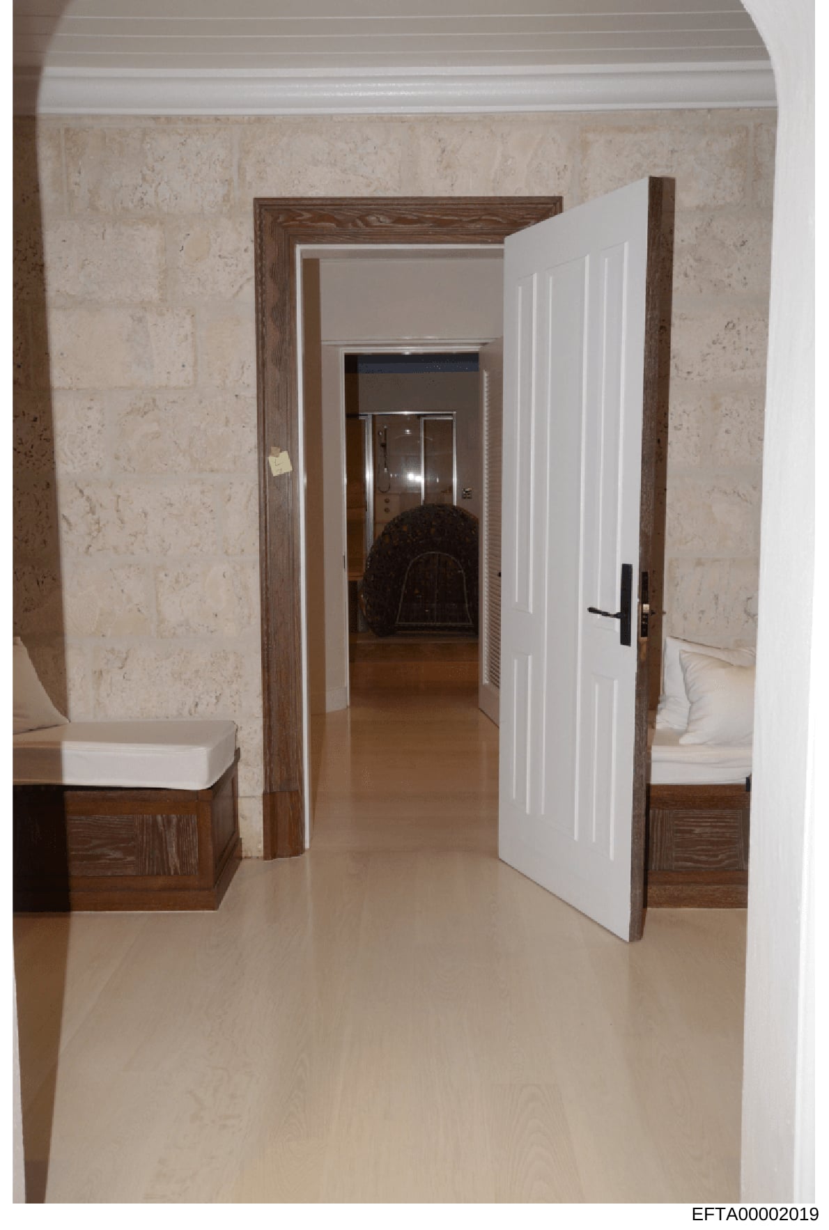 Photograph of an interior hallway featuring marble-clad walls, dark wood door frames, white paneled doors, and polished flooring. The image shows a series of connected rooms with built-in wooden benches or storage on either side. This appears to be d