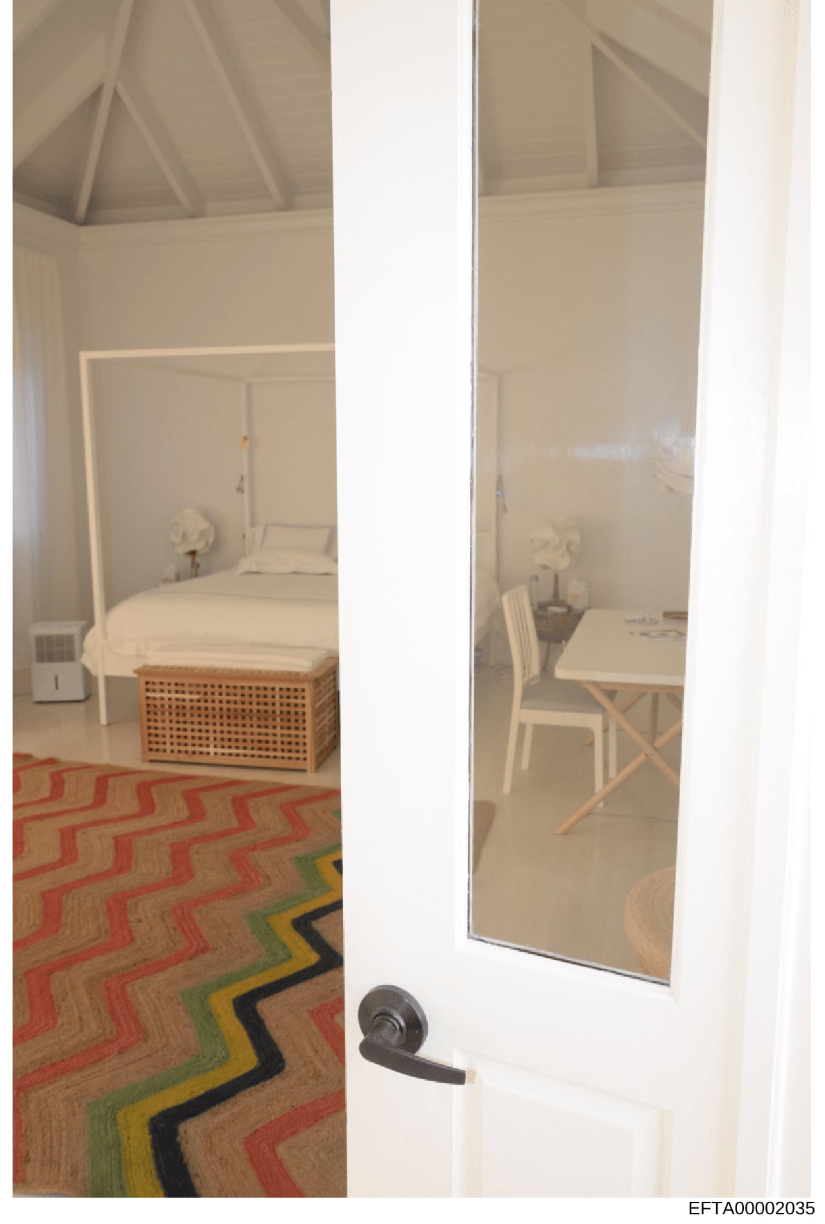 This is an evidence photograph showing the interior of a room with a bedroom area visible on the left and a workspace on the right, separated by an open doorway. The image captures furnishings including a bed with white bedding, a wooden storage benc