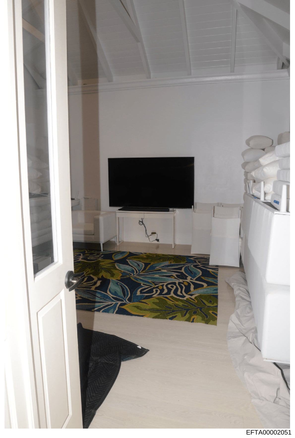This is a photograph of an interior bedroom space, showing a flat-screen television mounted on a white media console, contemporary furnishings, and decorative elements. The room features white walls, a patterned area rug with blue, yellow, and teal c