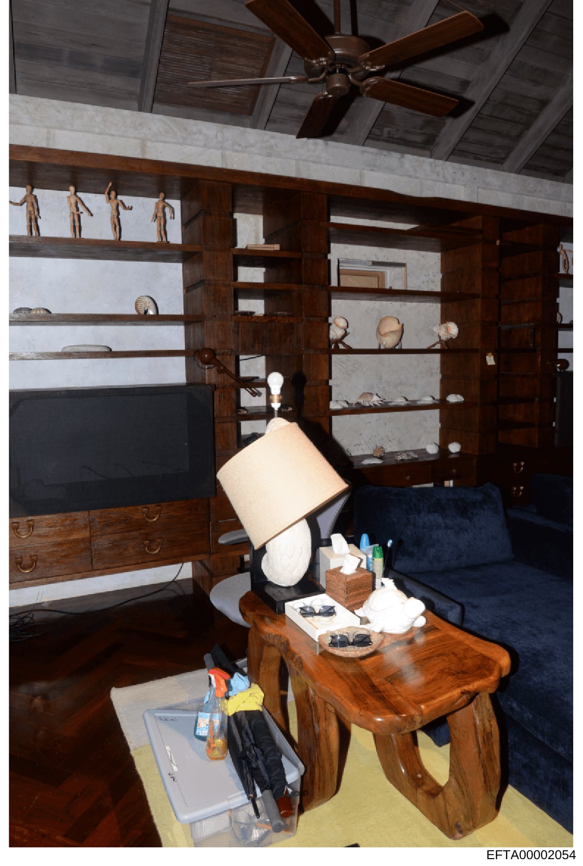 This is an evidence photograph showing the interior of a residential room, likely from one of Epstein's properties. The image captures a contemporary living space with high-end furnishings including a flat-screen TV, custom wooden shelving units disp