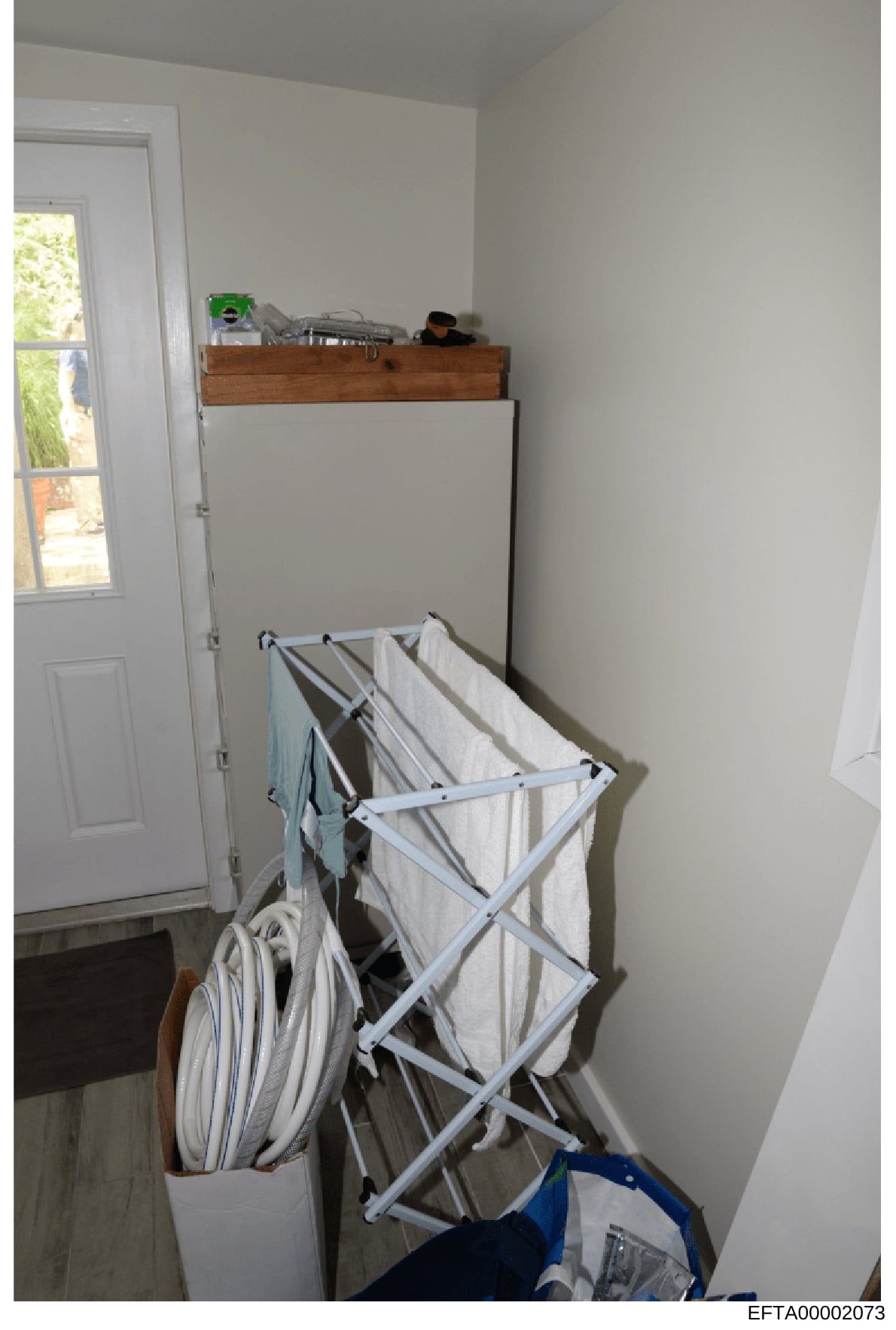 This is a photograph of a utility room or laundry area within a residential property. The image shows a white-painted room with a collapsible drying rack holding white linens/towels, a wooden dresser or cabinet with various items on top, and a door w