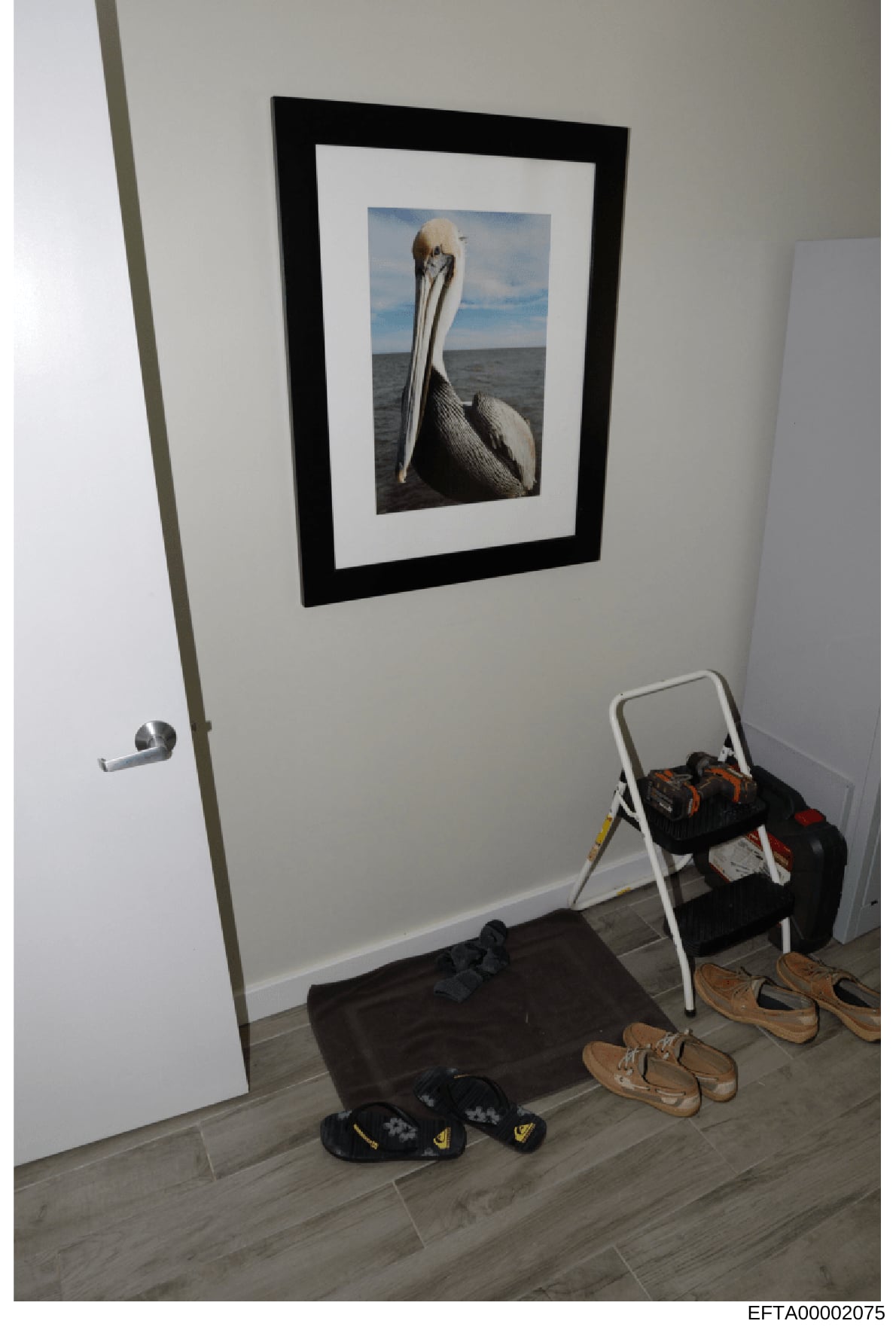 This is a photograph of an interior room space showing a framed pelican photograph hung on a white wall, with various personal items including shoes, a stepladder with clothing, and other belongings on the floor. The image appears to be evidence docu