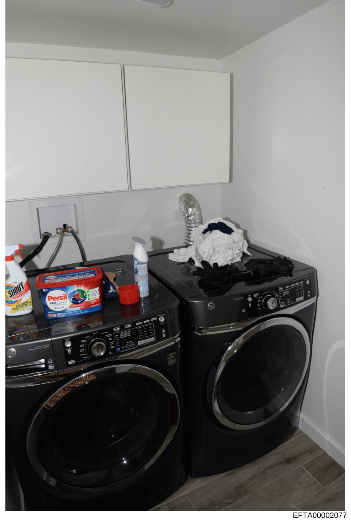 This is an evidence photograph showing a residential laundry room containing two black front-load washing machines, cleaning supplies, and miscellaneous household items. The photo appears to be part of property documentation in the investigation, cap