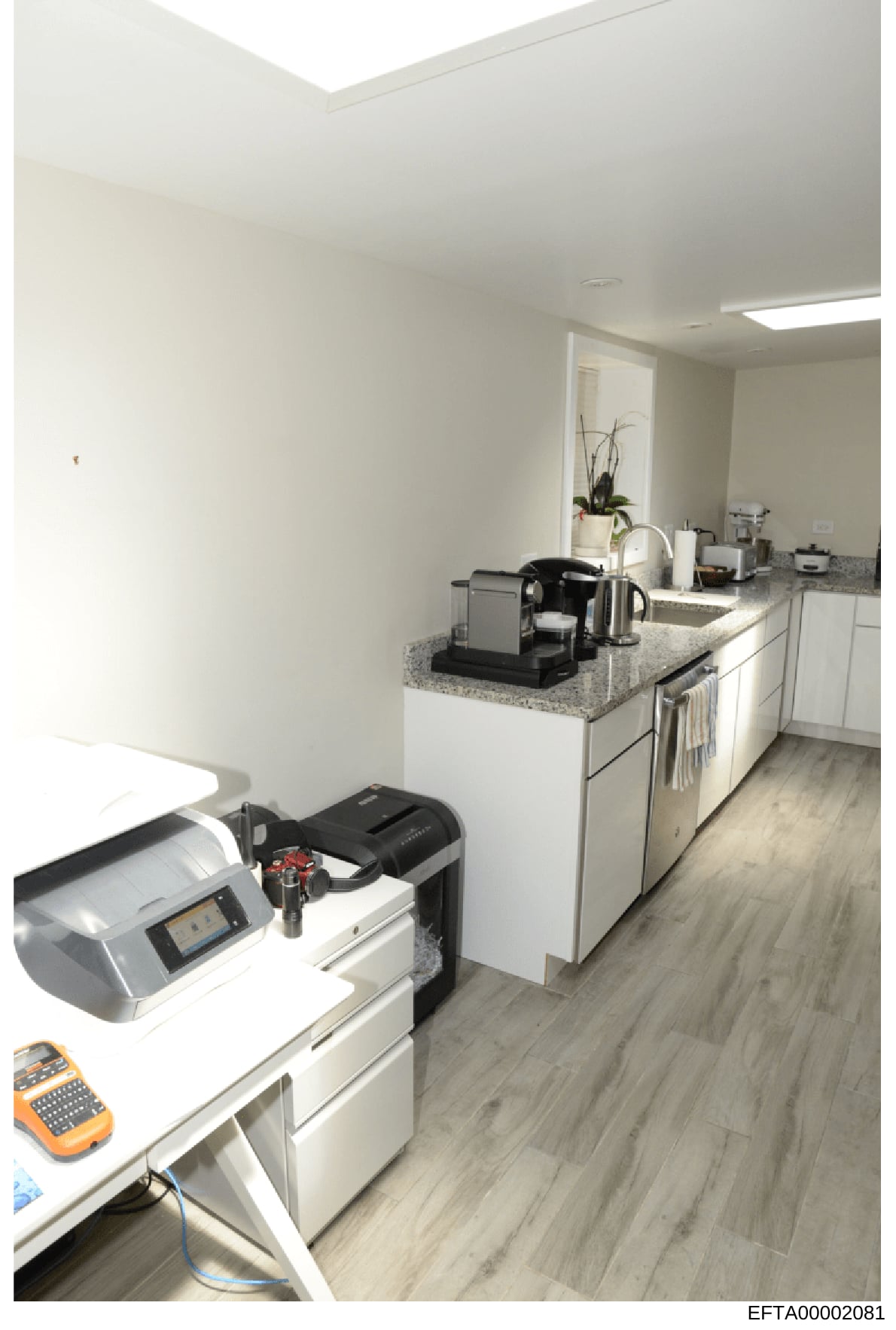 This photograph shows a modern kitchen space with contemporary cabinetry, granite countertops, and integrated appliances. The kitchen also contains office equipment including a label maker and other devices, suggesting this space served dual purposes