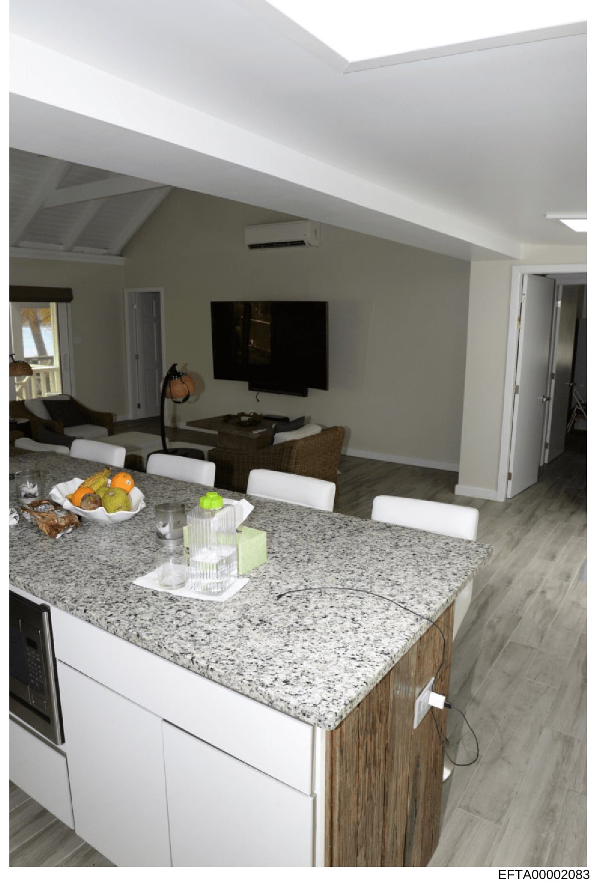 This is a photograph of a contemporary kitchen and adjoining living area, showing a granite-countertop island with white cabinetry, stainless steel appliances, and an open-concept layout with a media room visible in the background. The photo appears