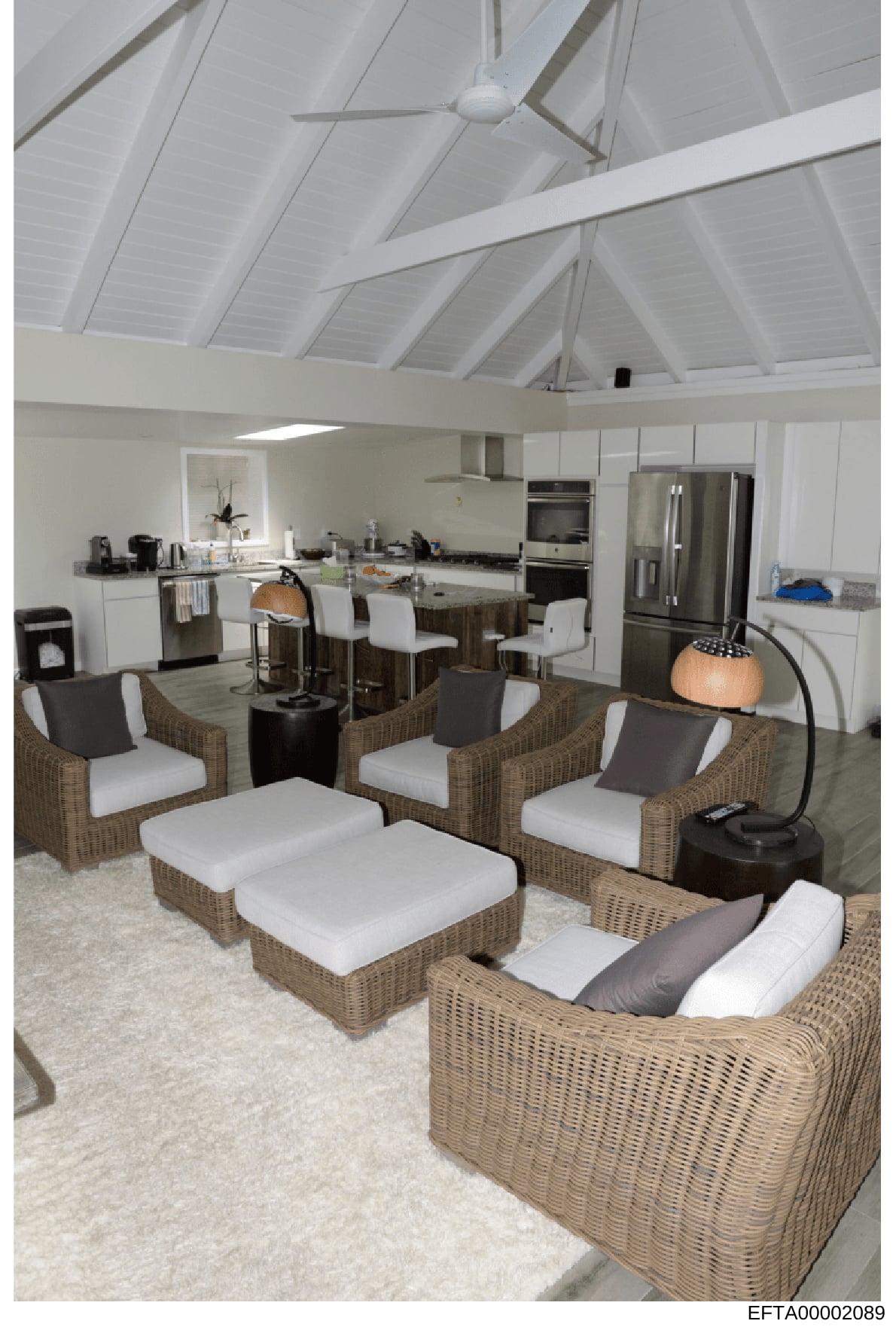 This is an interior photograph showing a modern, light-filled residential space with an open-concept design combining a kitchen and living area. The image depicts high ceilings with exposed white beams, stainless steel appliances, wicker seating furn