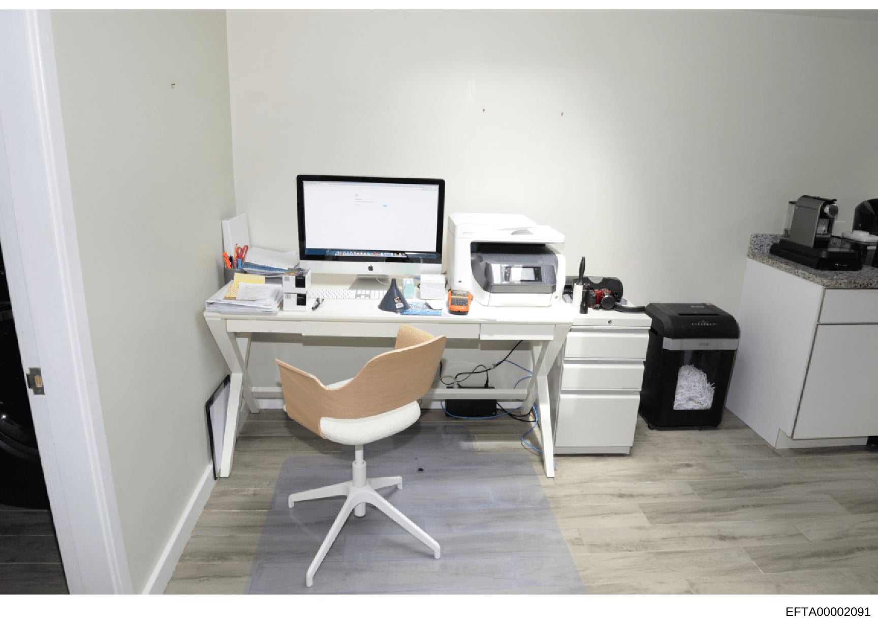 This is a photograph of a modern office or workspace interior, featuring a white desk setup with an iMac computer, printer, office supplies, and a wood-and-white ergonomic chair. The room has light gray walls, wood-grain flooring, and includes filing